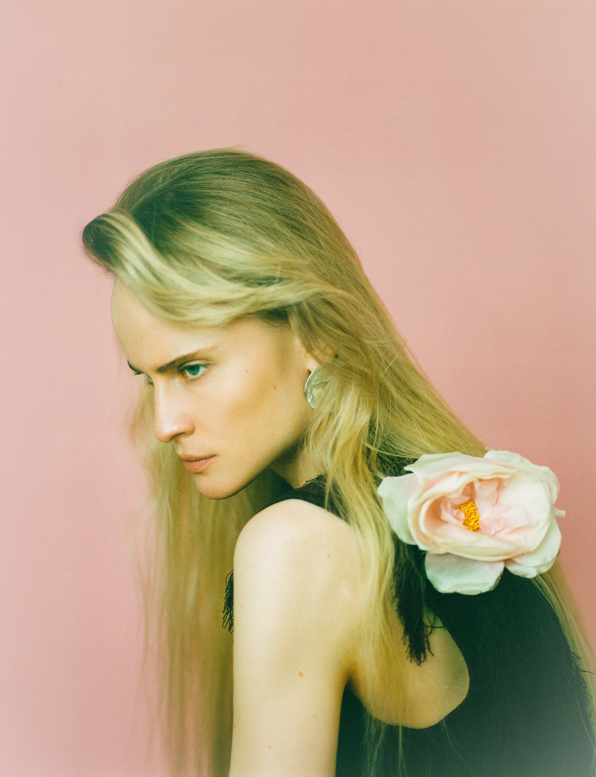 Blonde person with pink flower on shoulder, pink background.