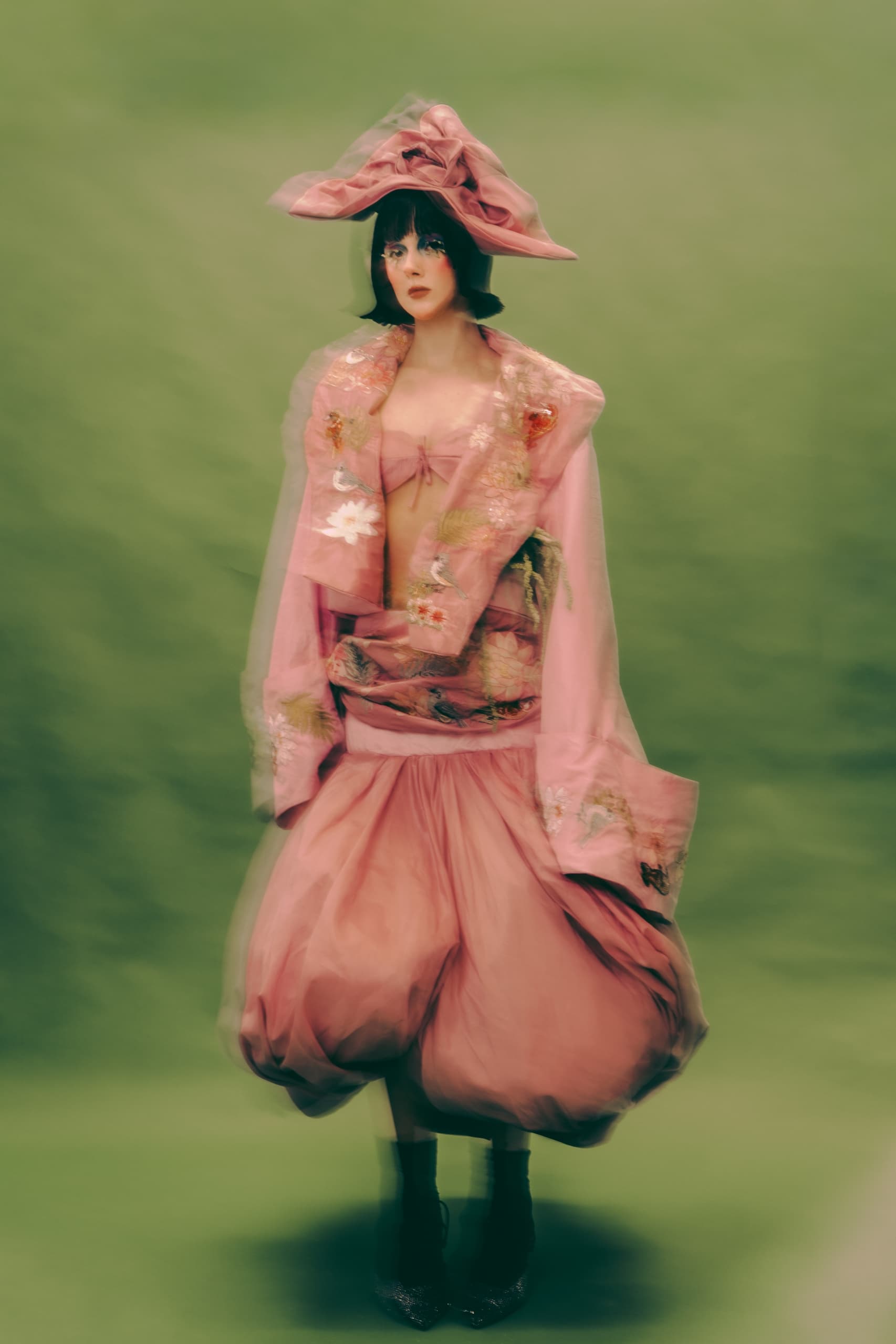 Model in pink avant-garde outfit with large hat on a green backdrop.