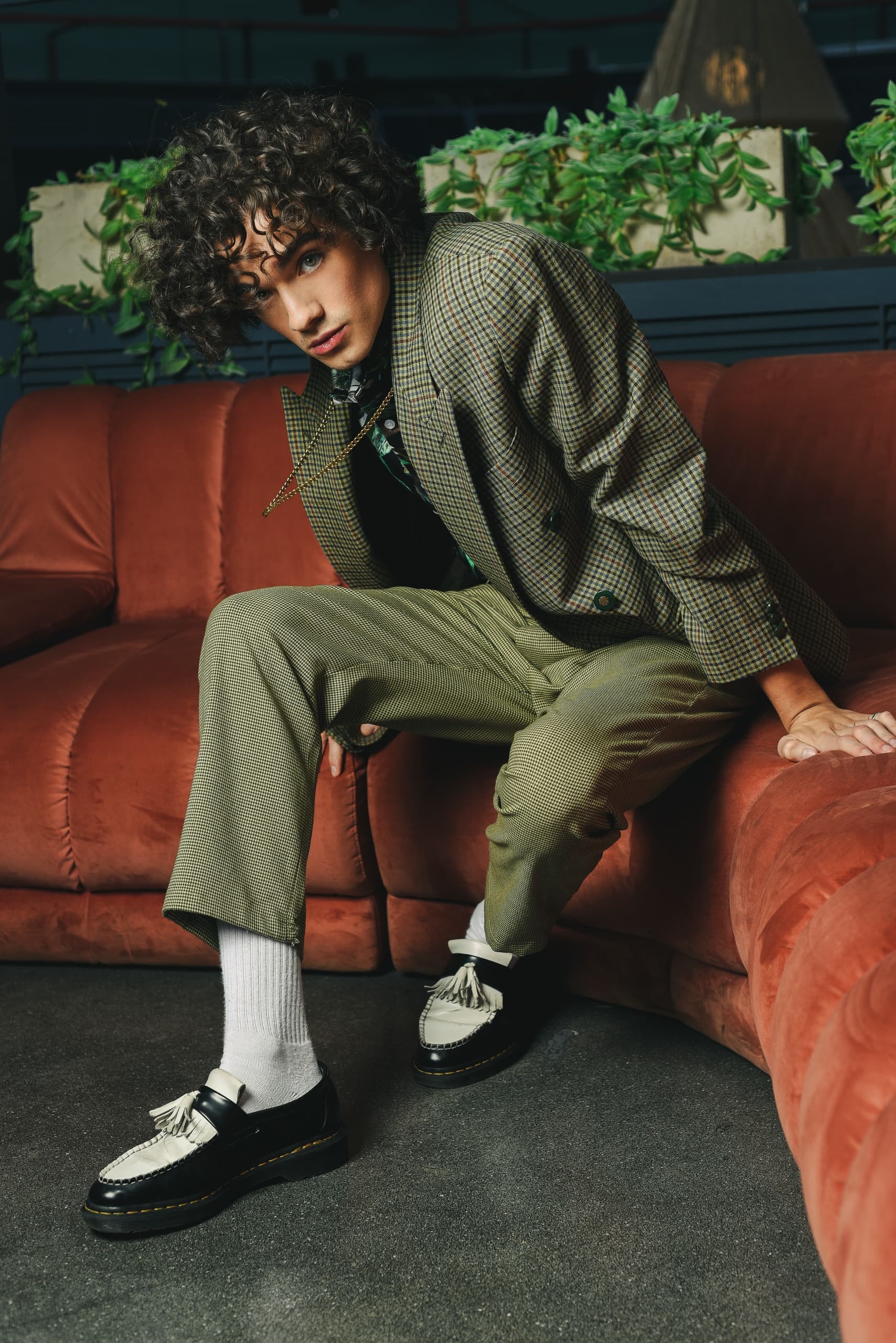 Fashionable person in checked suit on a red couch with greenery background.