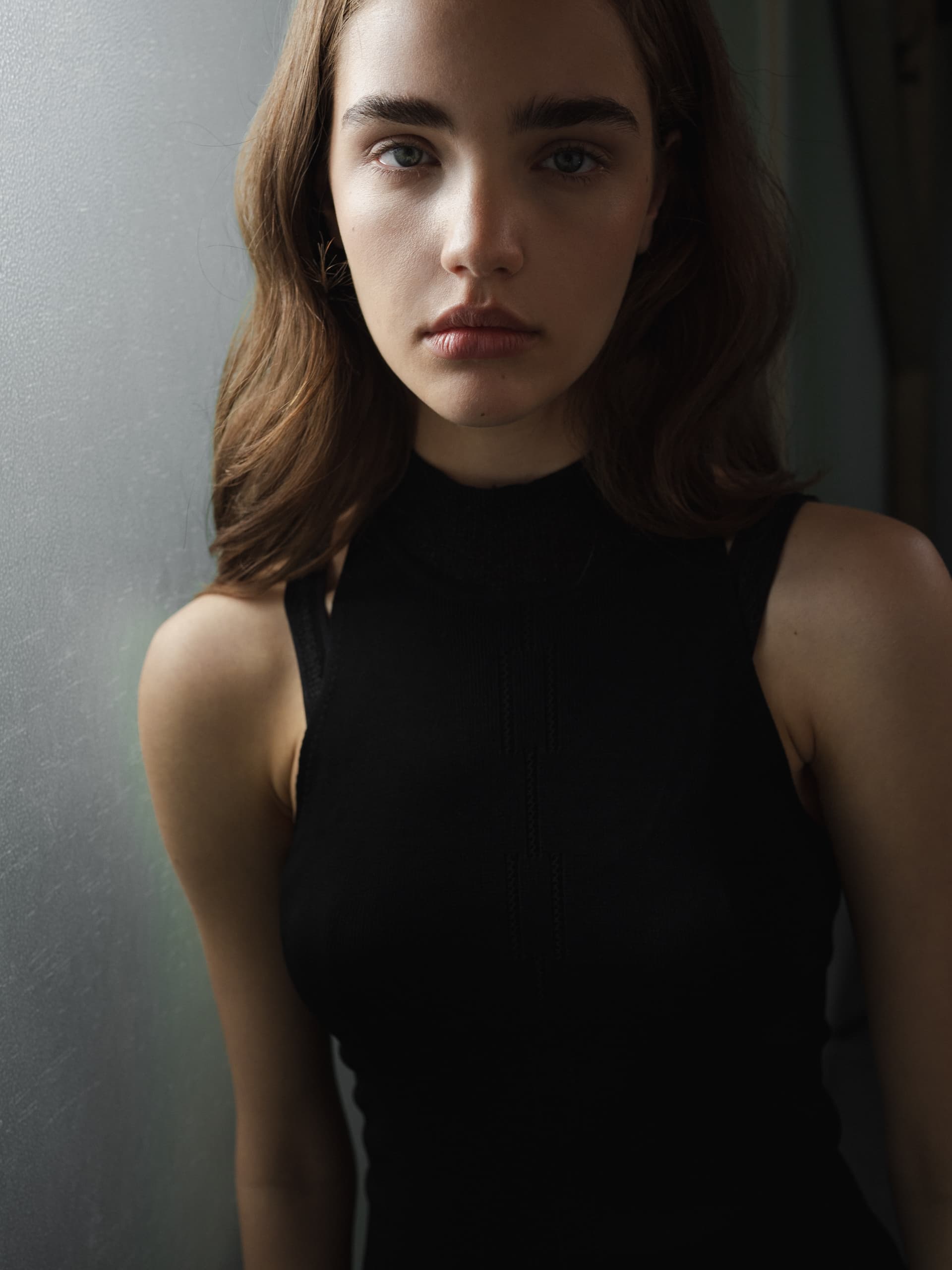Woman in black top with soft lighting, leaning against a textured wall.