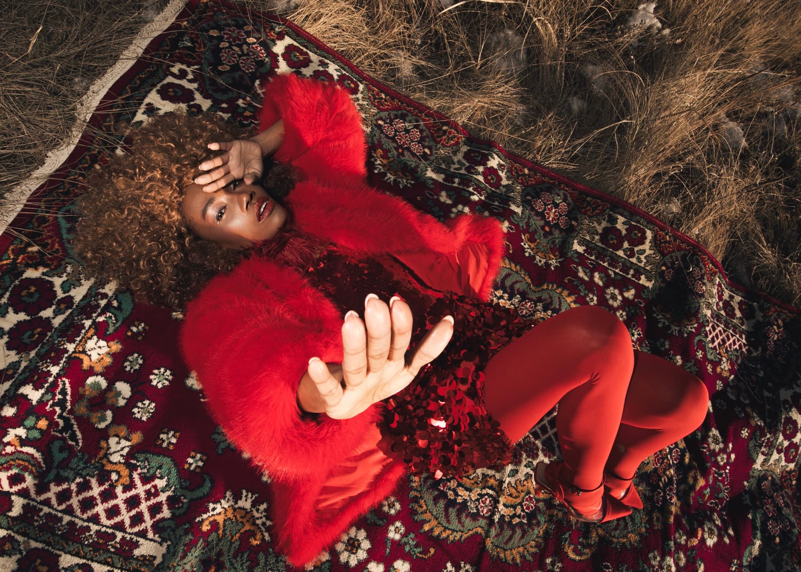 Fashionable person in red outfit lying on ornate rug outdoors.