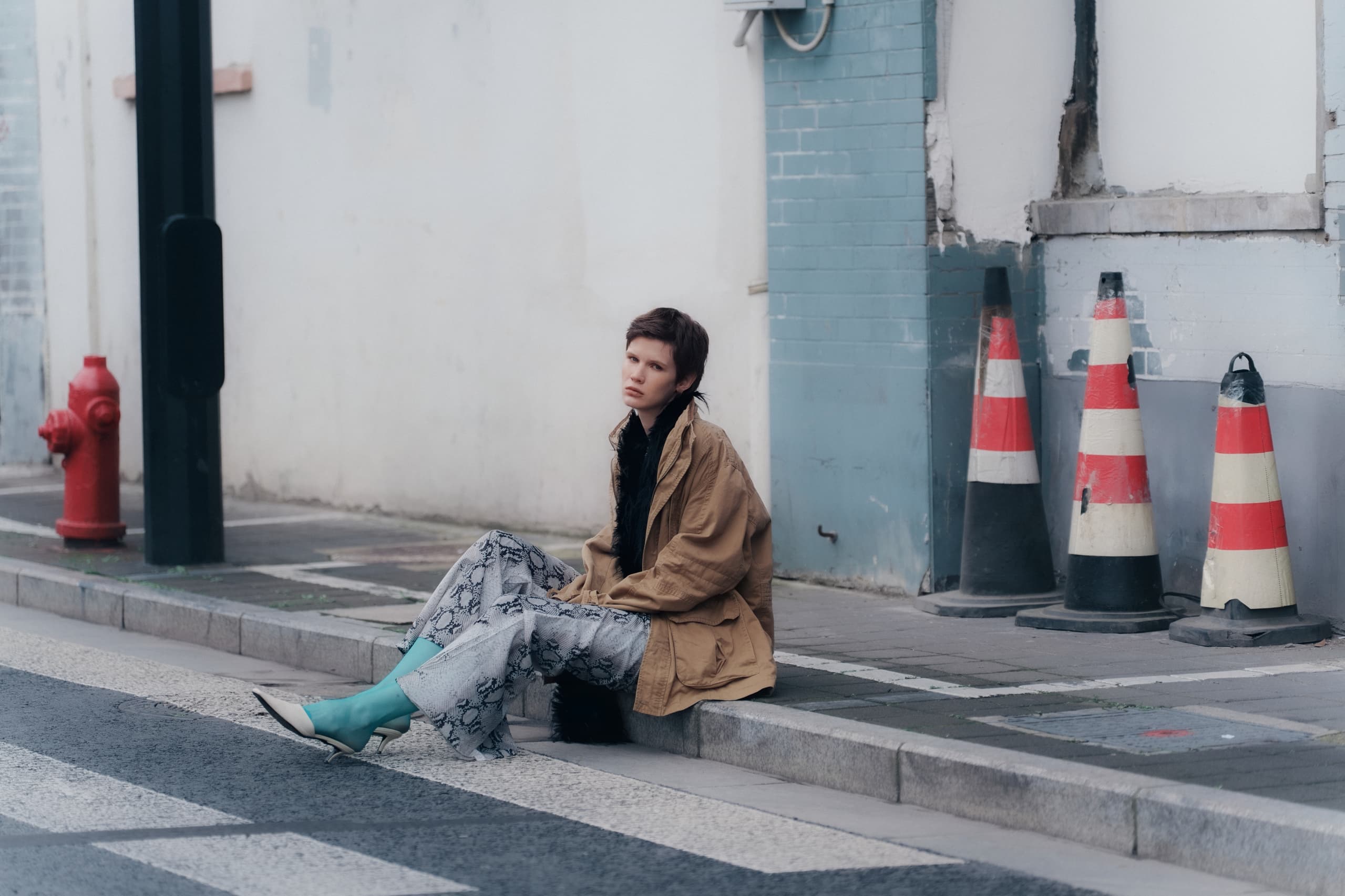 Person sitting on urban sidewalk near safety cones, wearing stylish outfit.