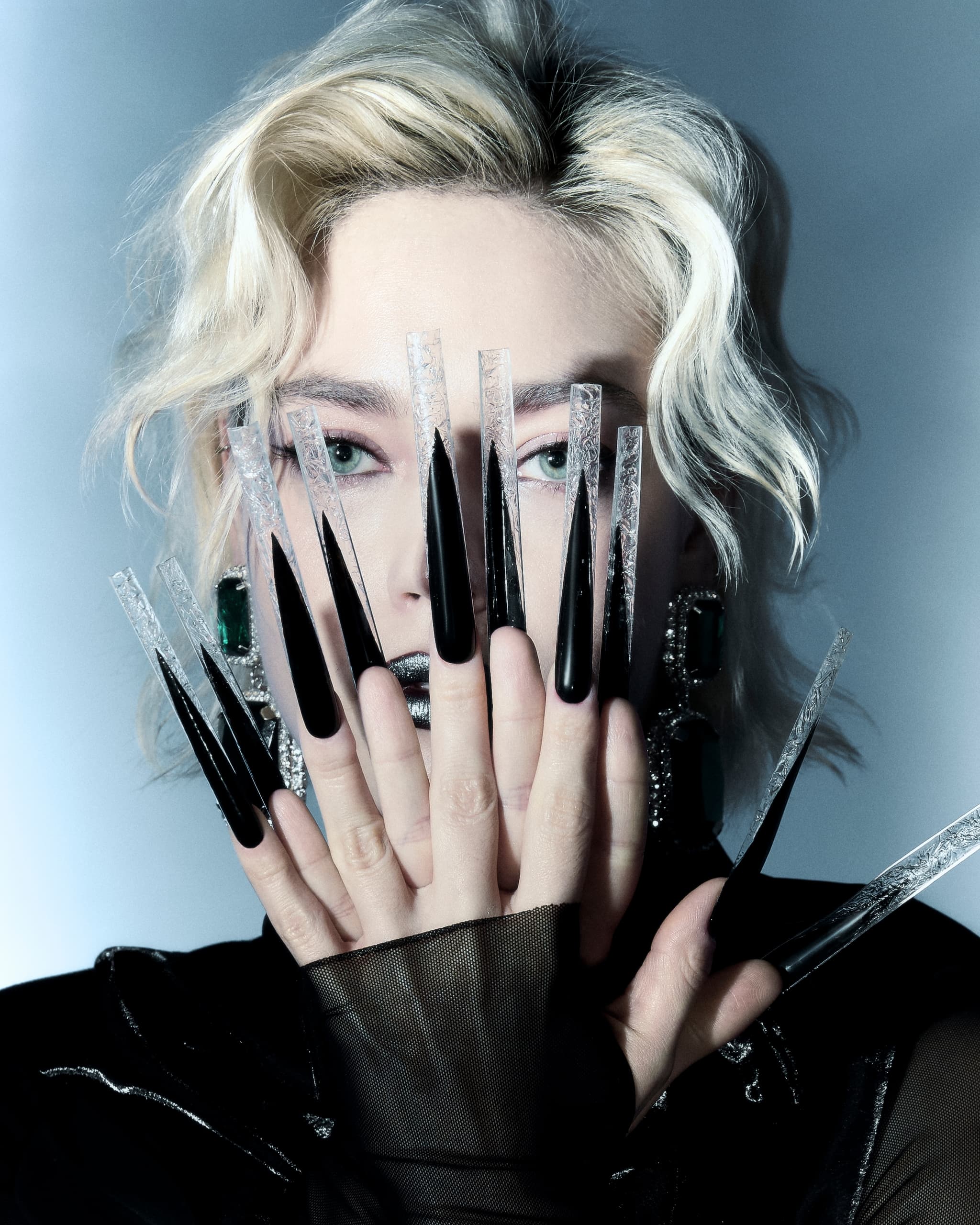 Blonde person with striking long black nails covers their face in dramatic style shot.