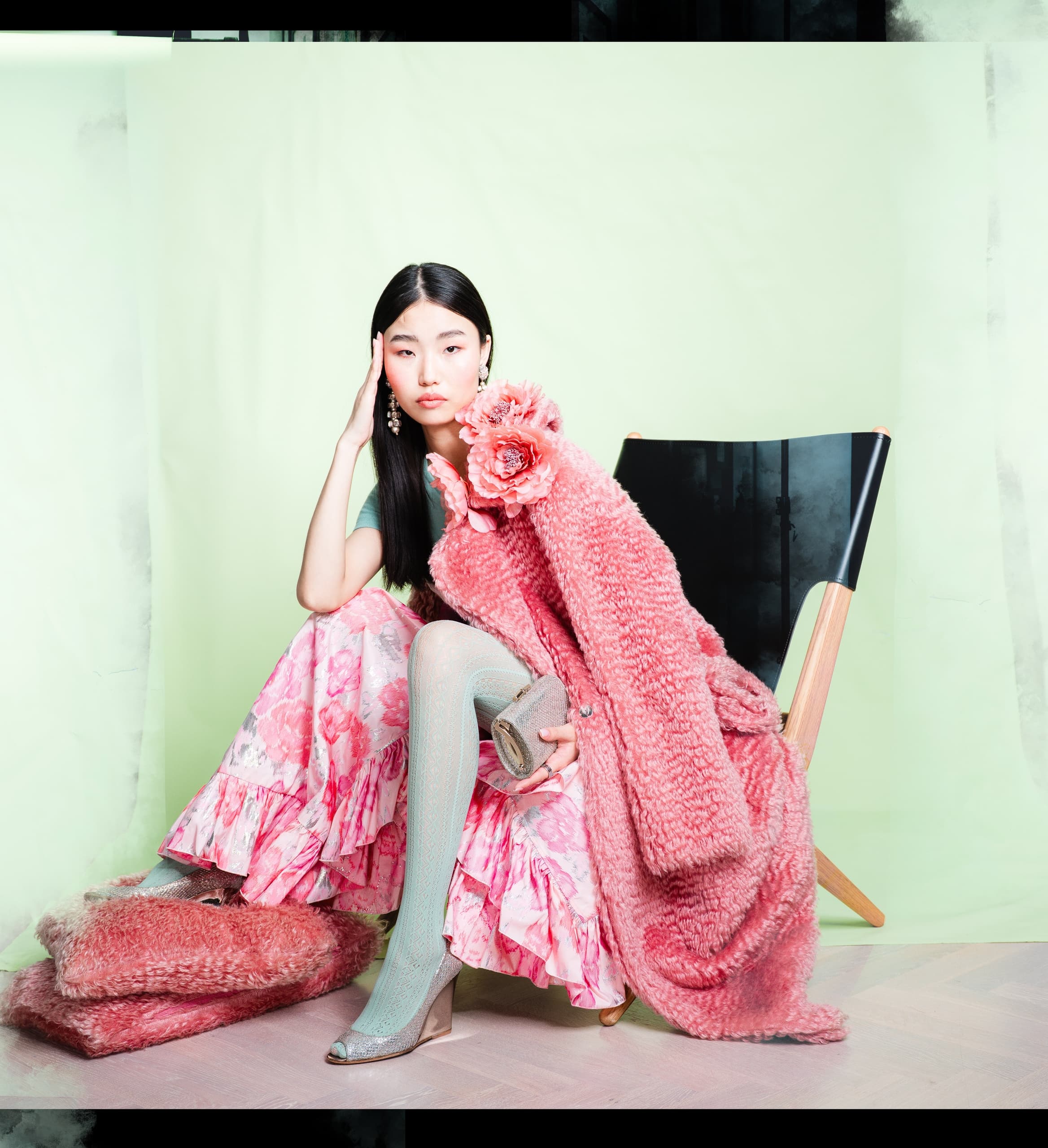 Stylish woman in floral dress and pink coat poses on chair against green backdrop.
