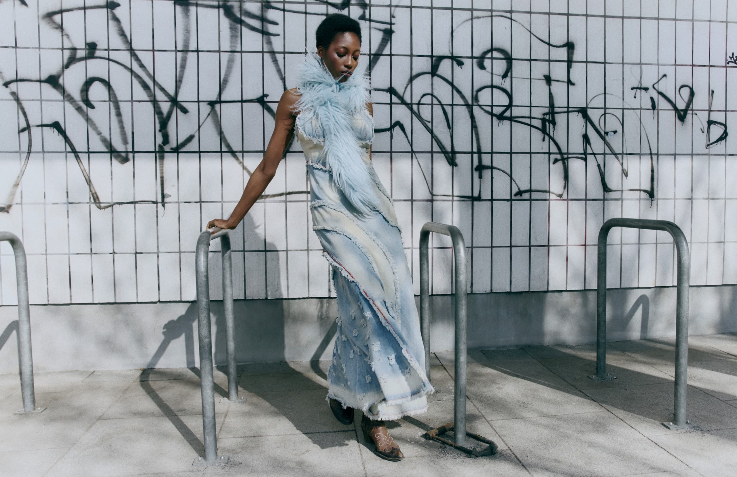 Person in a blue feathered dress leaning against a tiled, graffiti-covered wall.