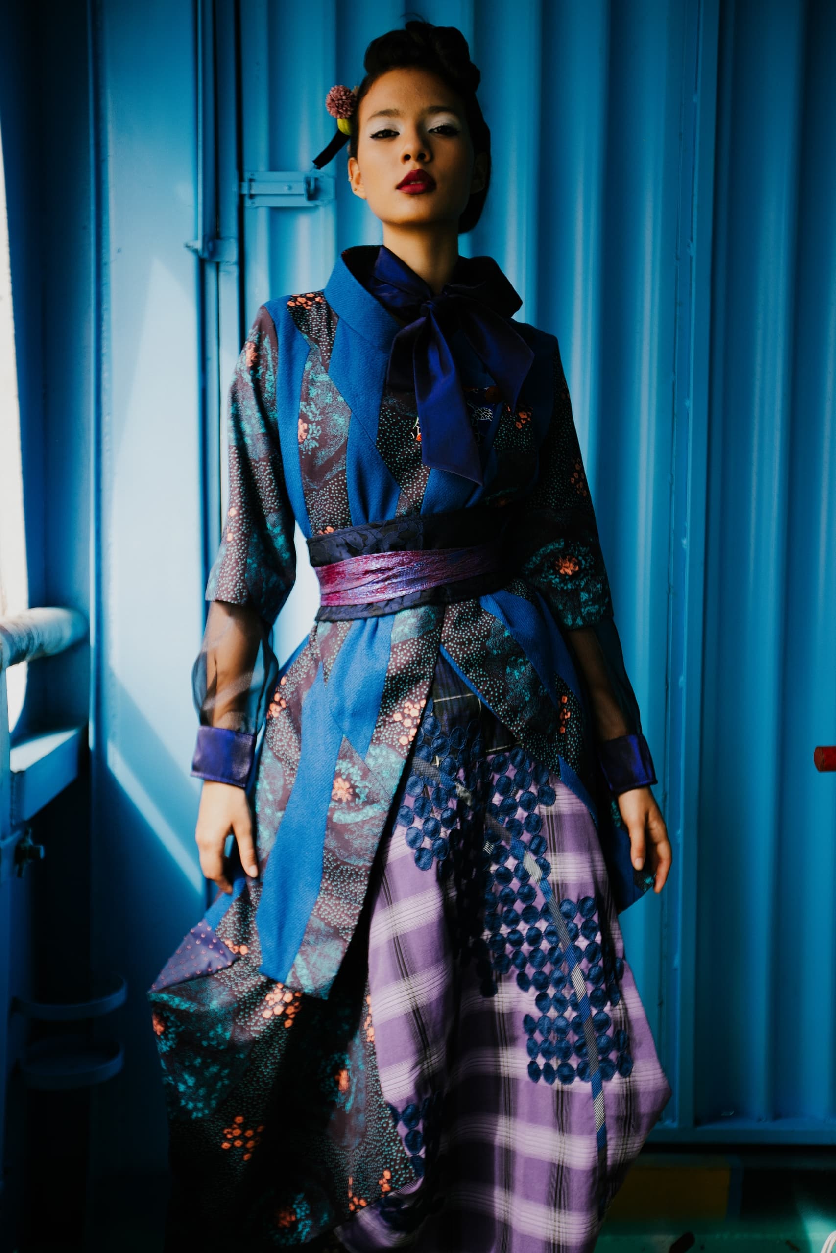 Fashionable woman in blue kimono-style outfit against blue wall.