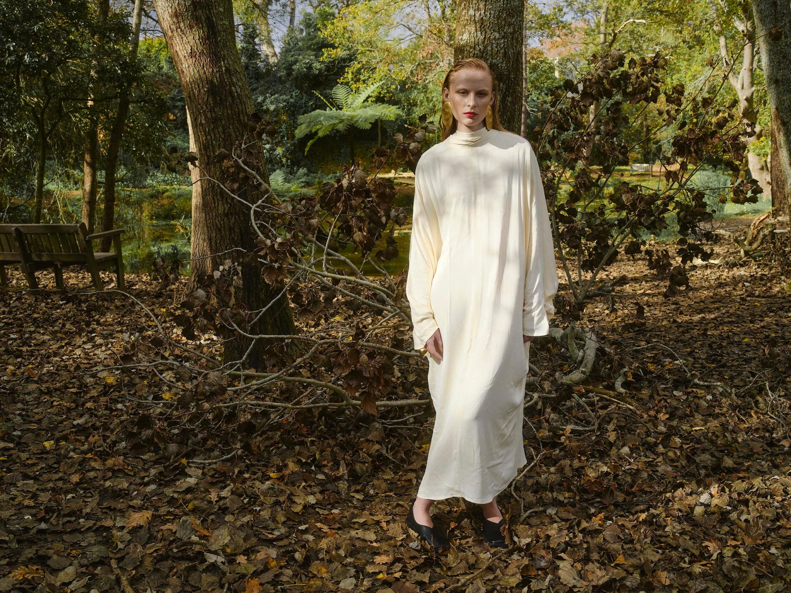 Woman in white dress stands in a wooded, leafy autumn scene.