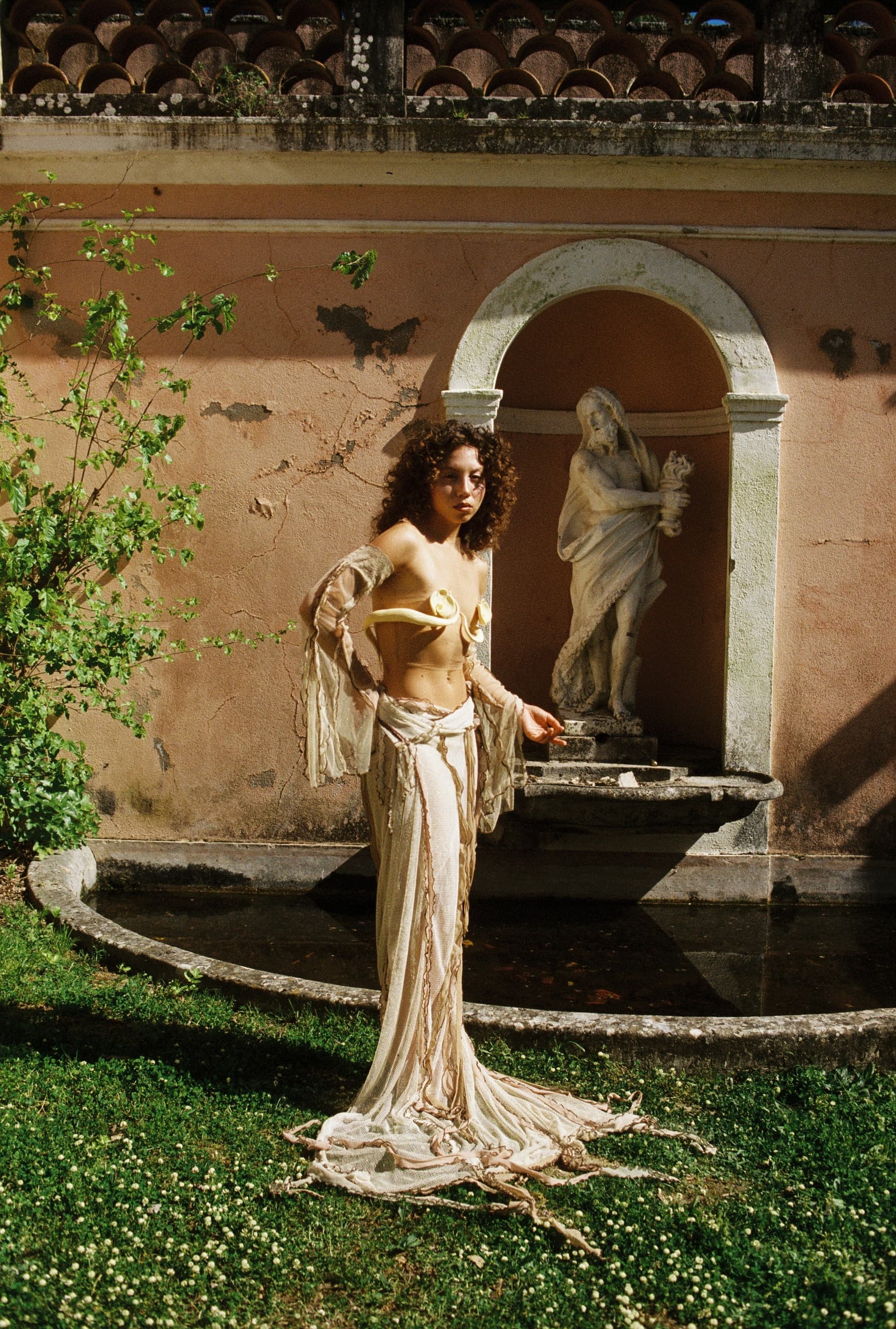 Woman in elegant attire by a garden statue fountain under sunny skies.