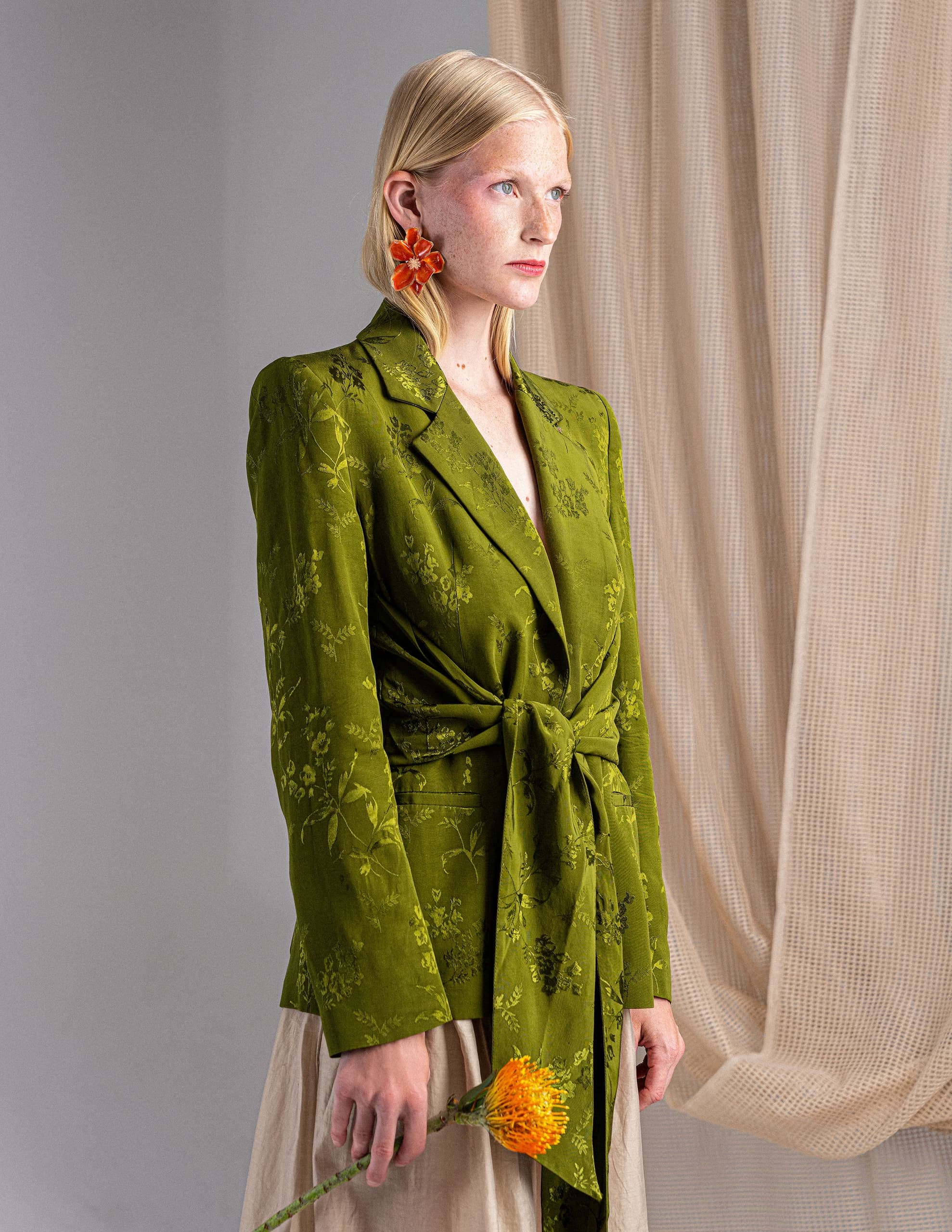 Woman in green floral blazer holding an orange flower, beige curtain background.