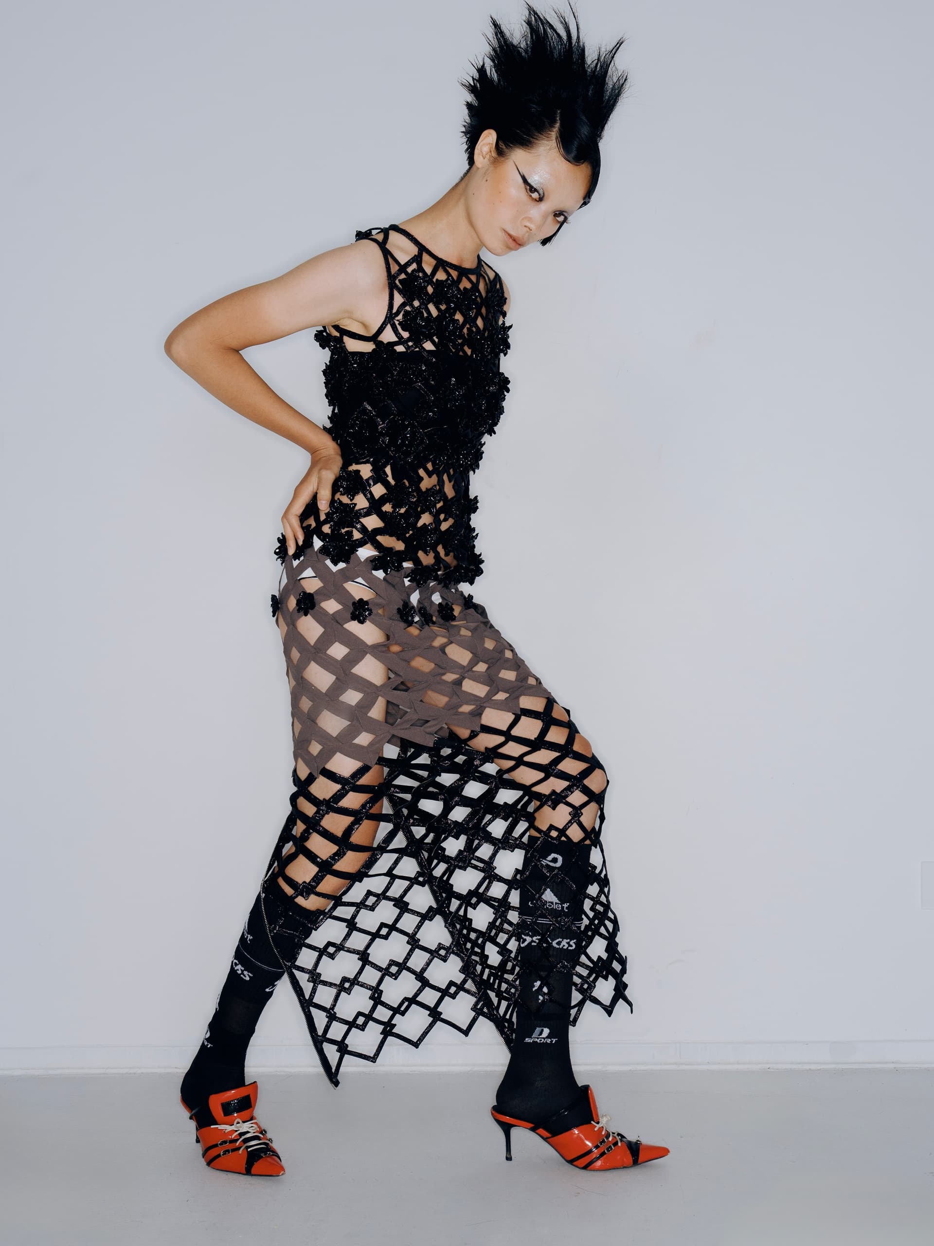 Stylish model in edgy black net dress and orange heels posing confidently.