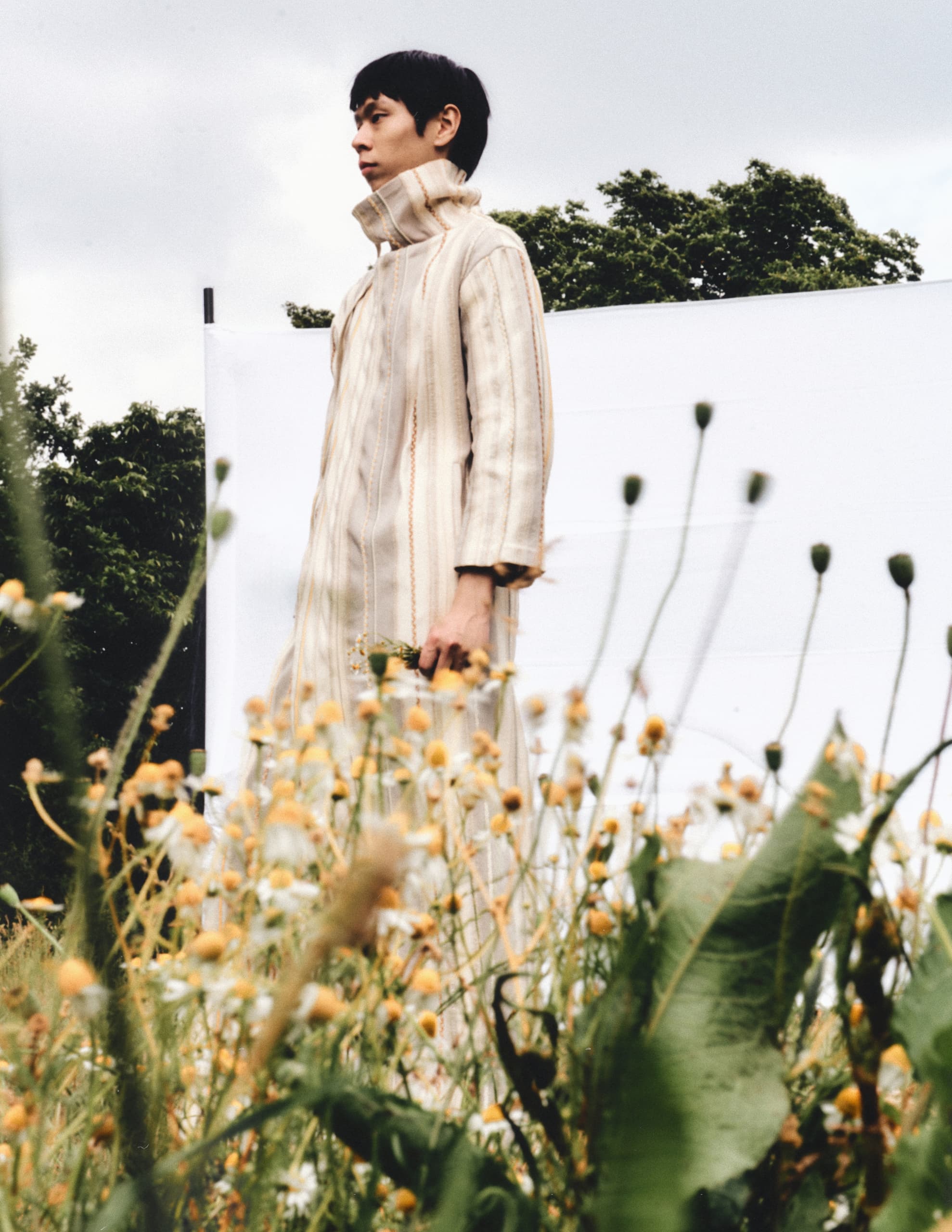 Person in a striped coat stands in a field of wildflowers under a cloudy sky.
