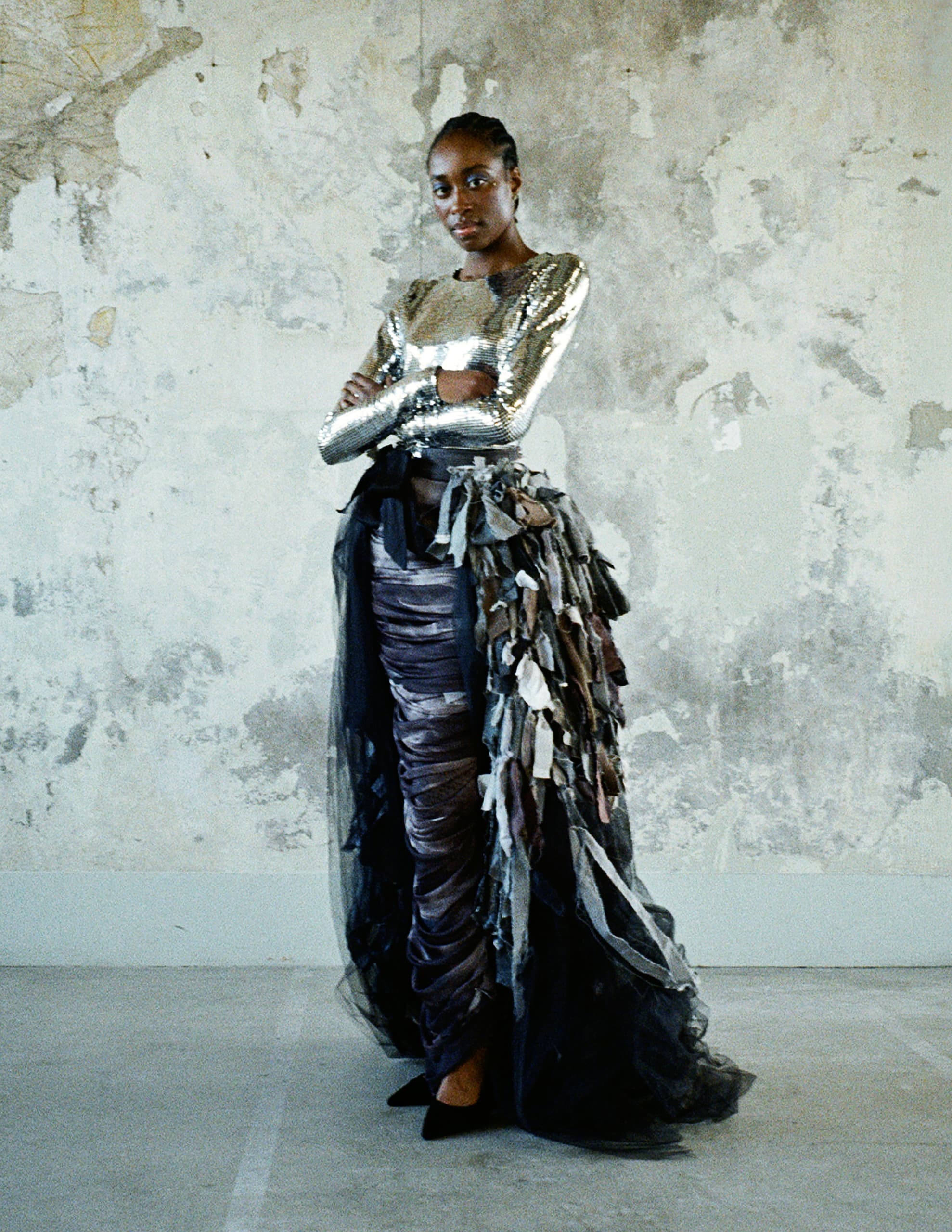Fashionable woman in silver top and layered skirt against textured wall.
