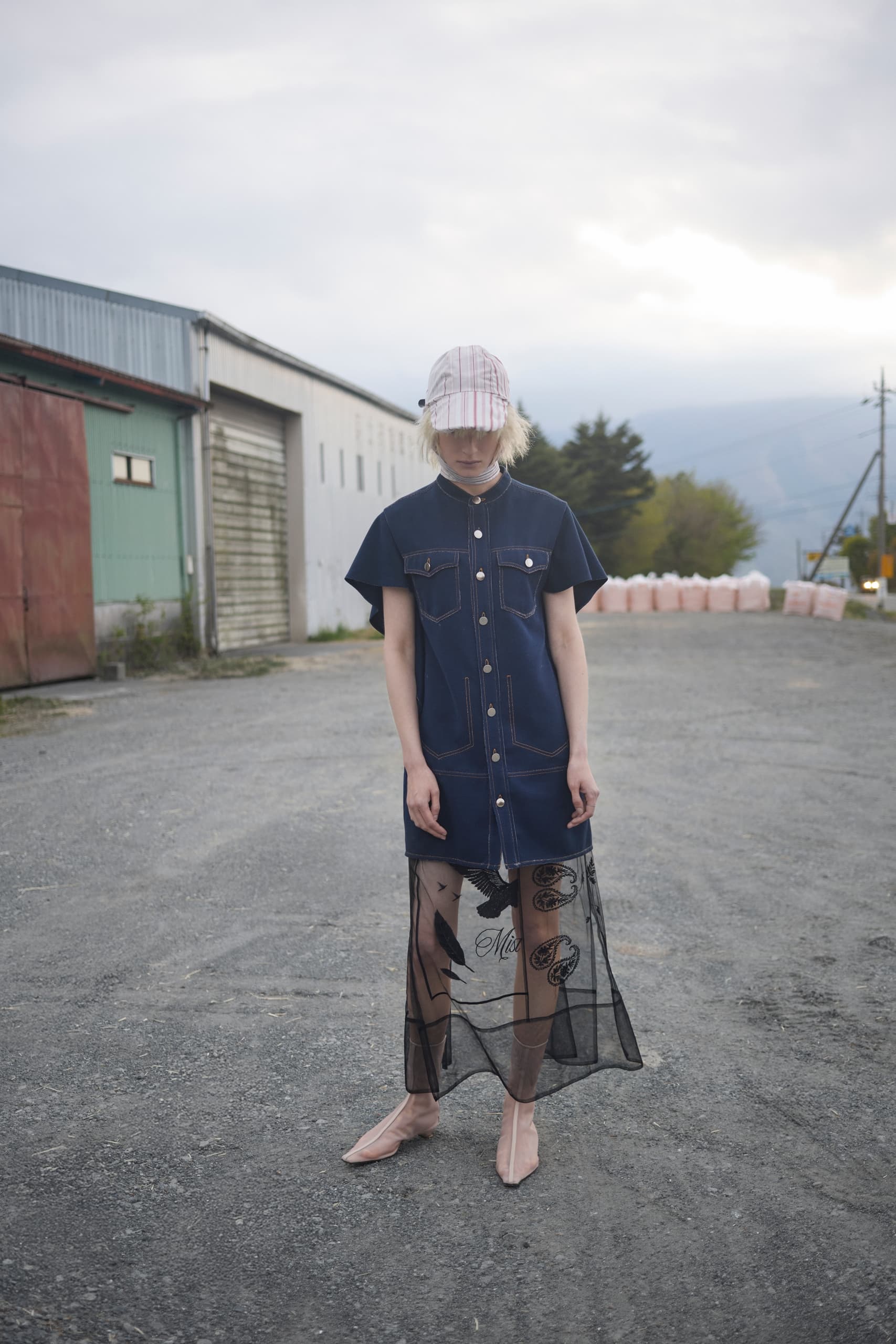 Fashionable person in denim dress and sheer skirt standing outdoors with cloudy sky.