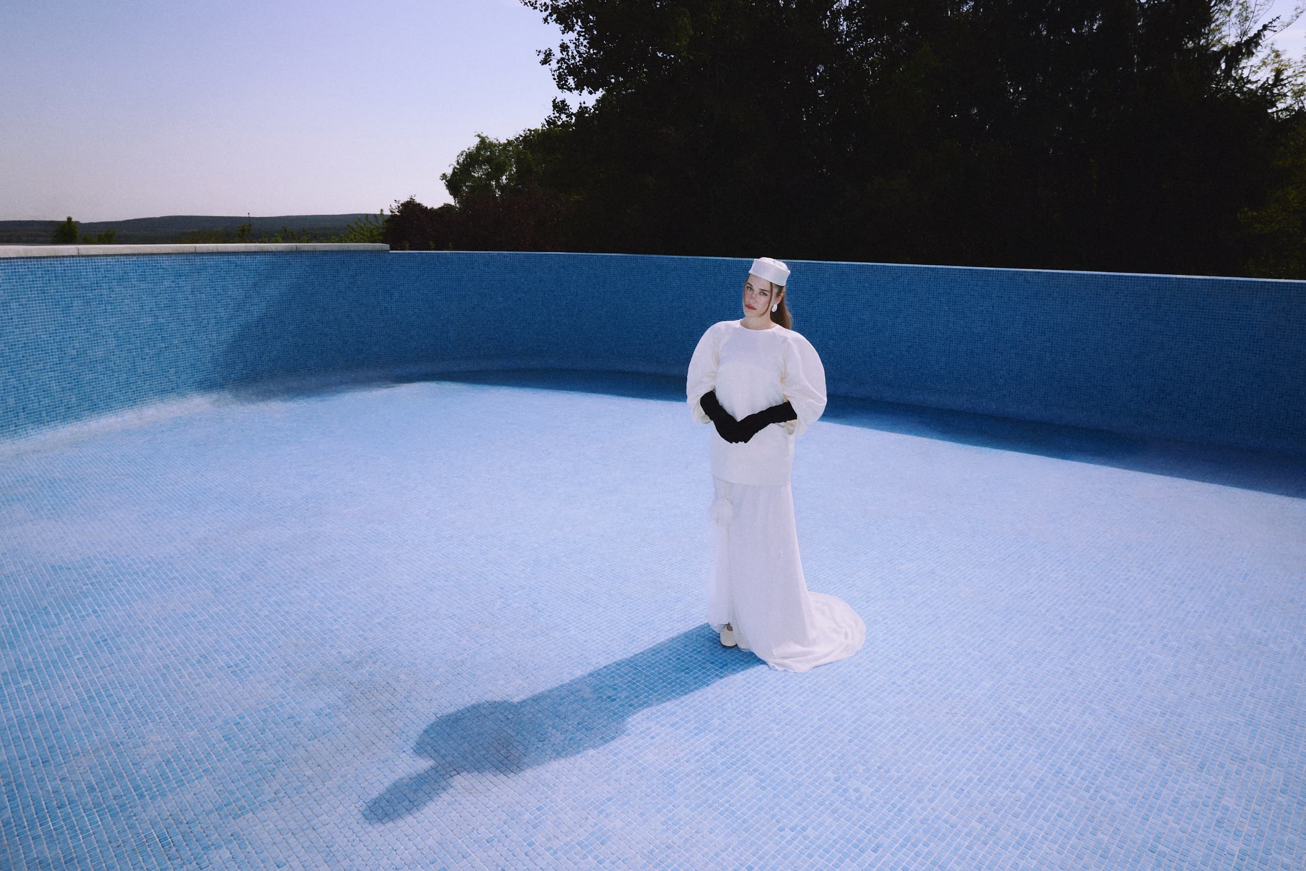 Woman in a white dress standing in an empty blue-tiled pool under clear sky.