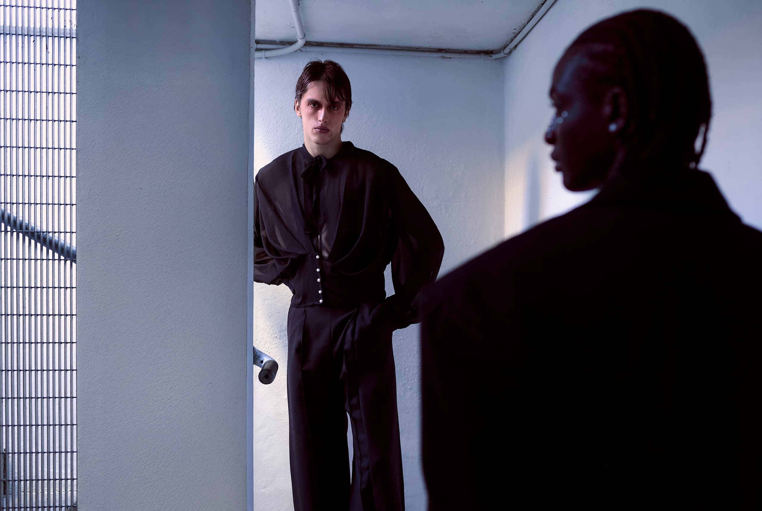 Two models in black outfits stand in a minimalist, modern setting.