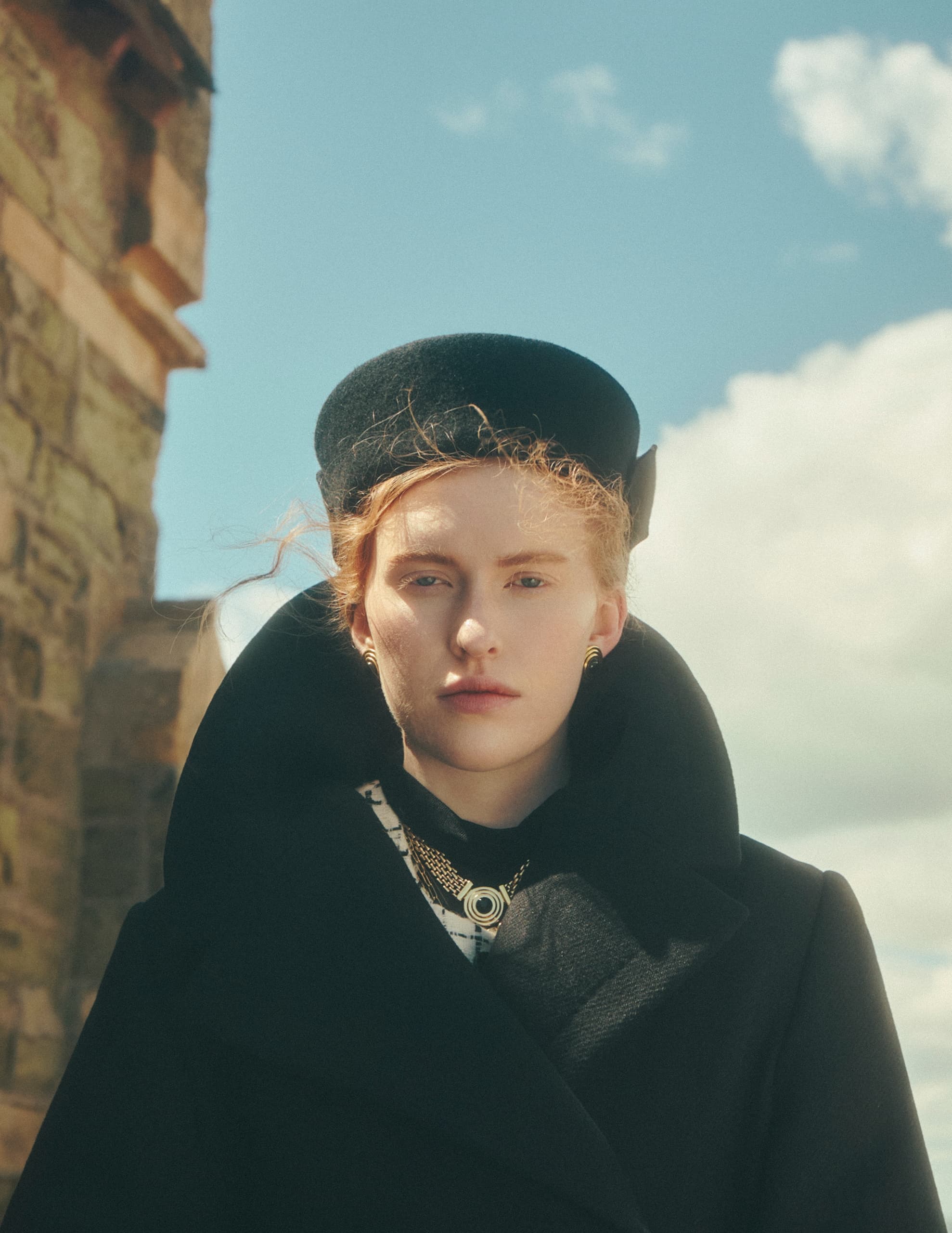 Woman in black coat and hat in front of old stone building under clear sky.