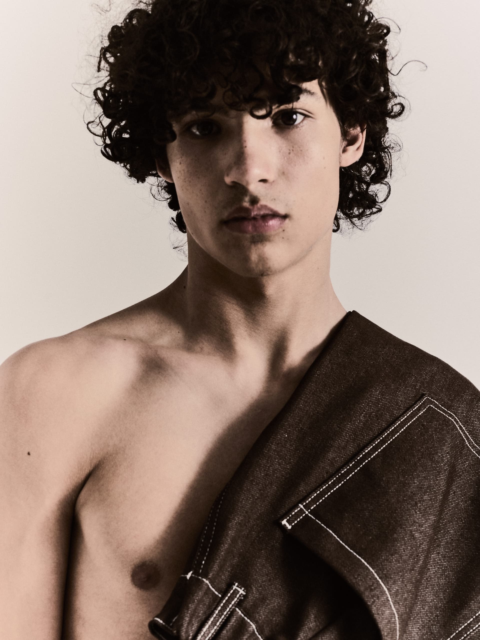 Young person with curly hair, bare shoulder, and fabric draped over, gazing at camera.