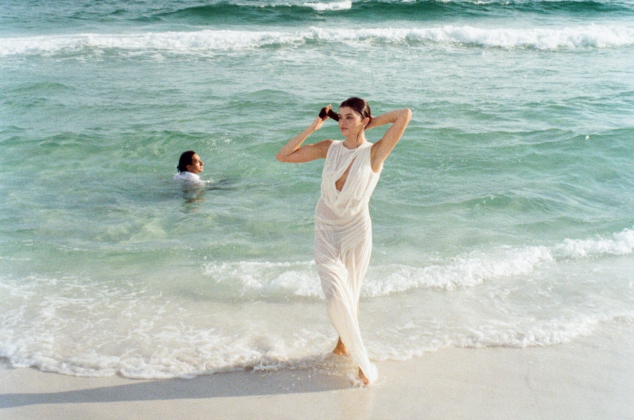 Woman in white dress walking on beach with ocean waves, summer getaway scene.