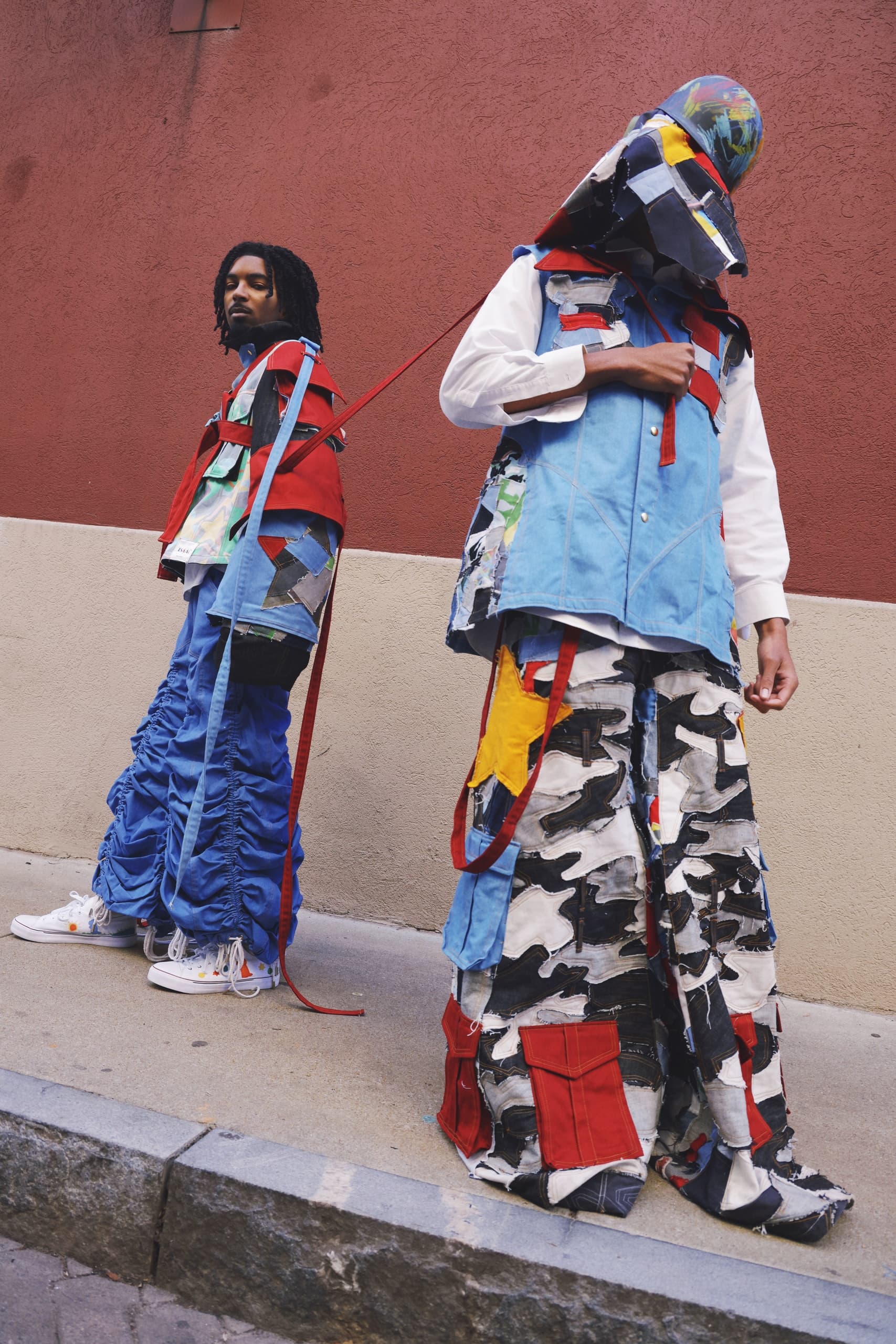 Two models in colorful patchwork outfits against red wall backdrop, showcasing street fashion.