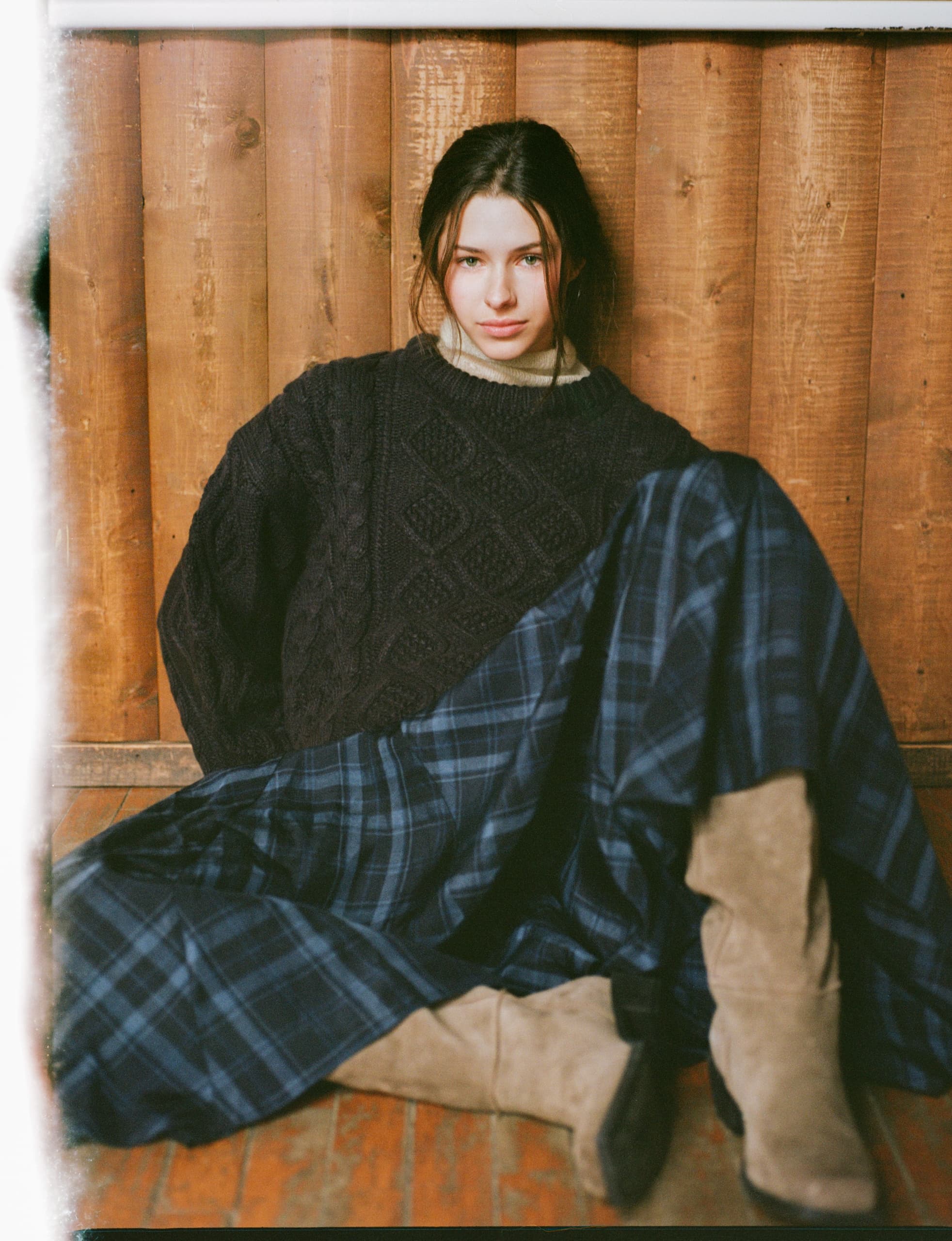 Woman in cozy knit sweater and plaid skirt, sitting against wooden wall.