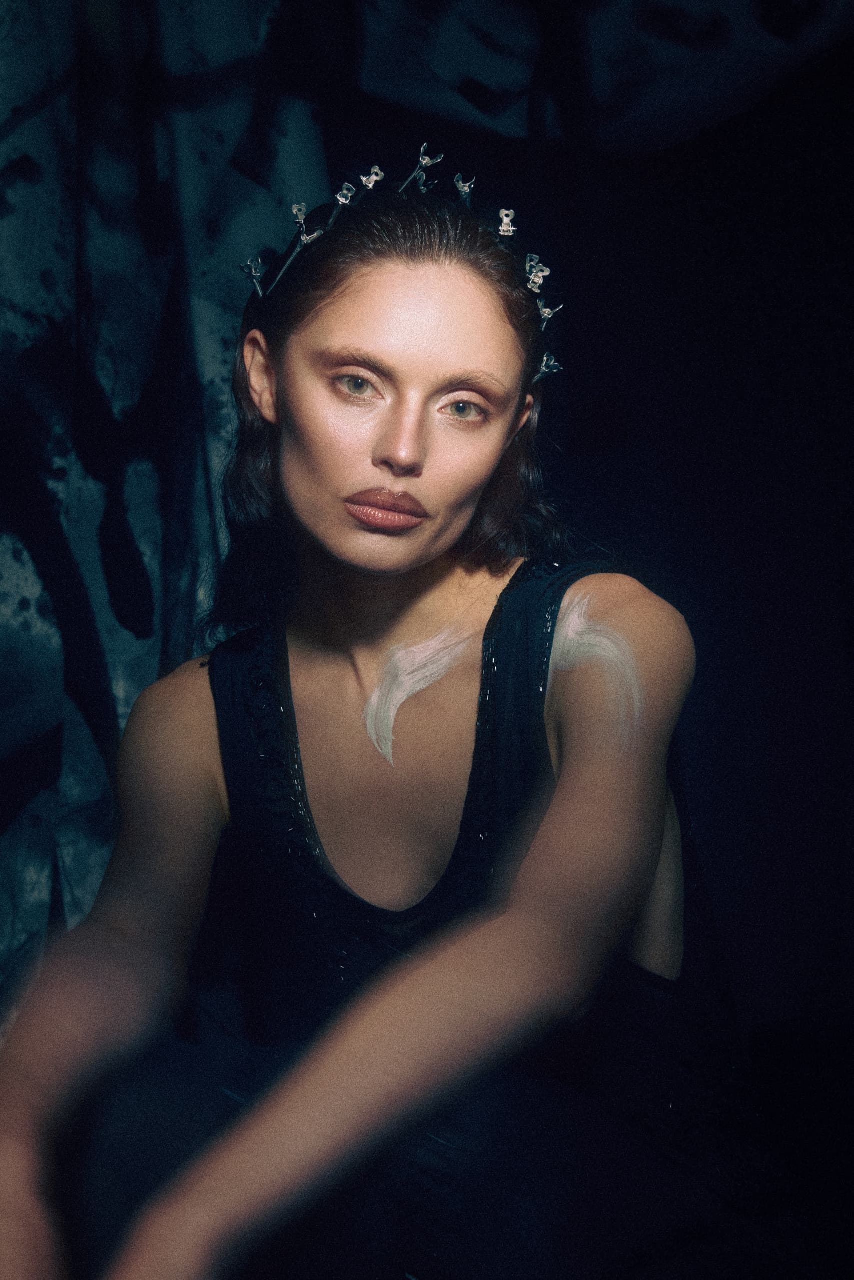 Woman with artistic expression sitting in dim light, wearing a decorative headpiece.