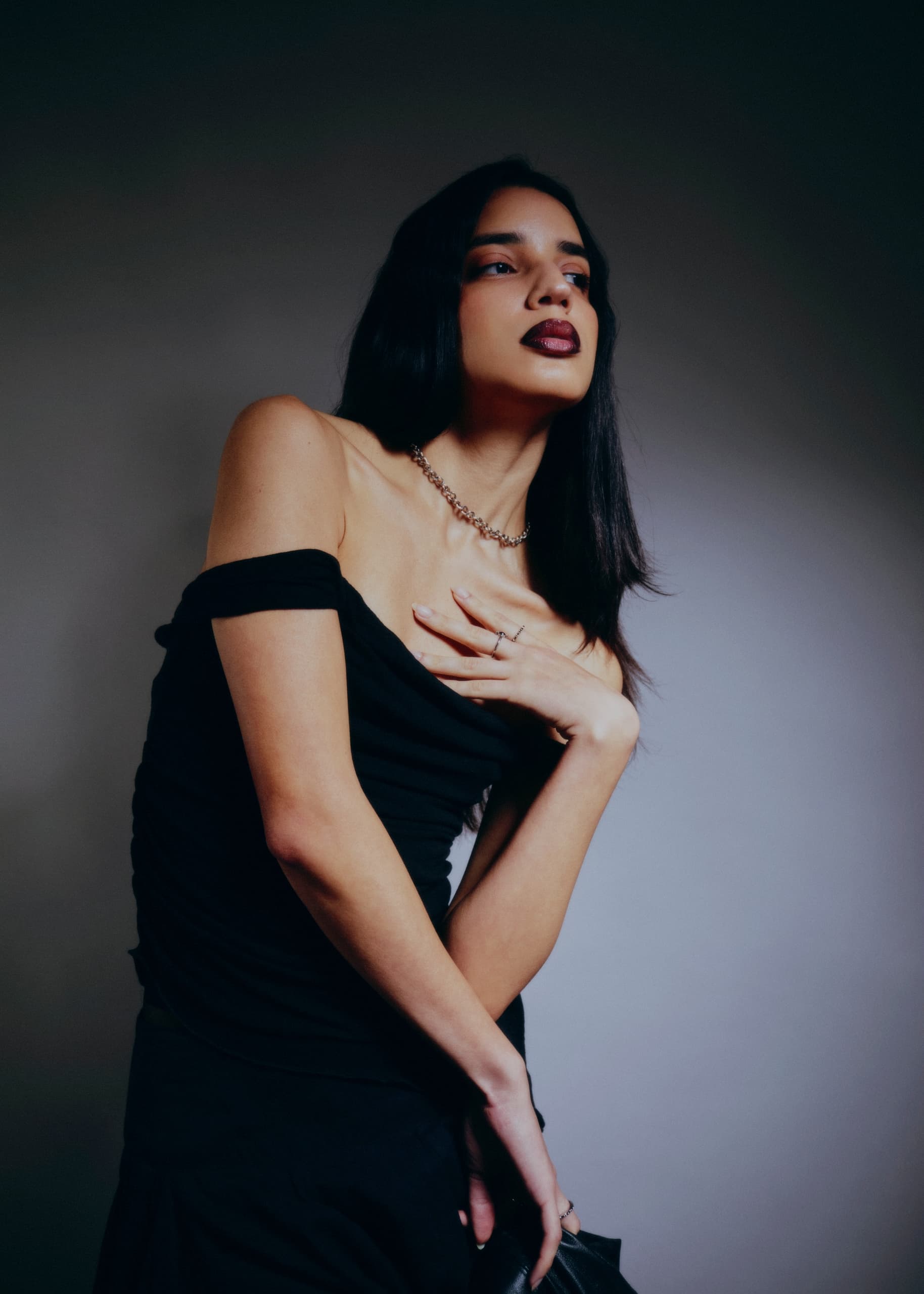 Fashion model in elegant black dress posing confidently in dimly lit background.