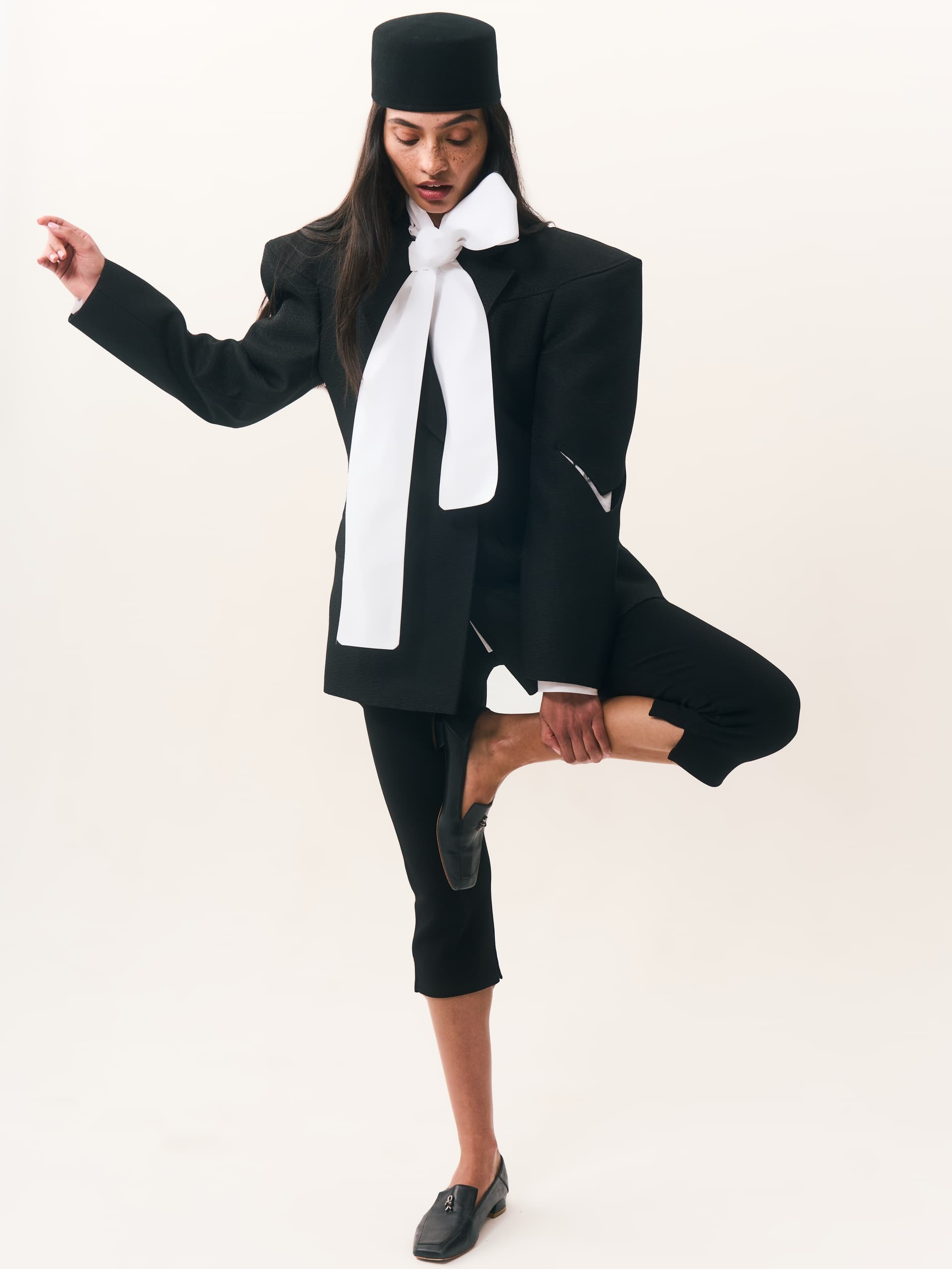 Fashion model in unique black suit with white scarf and hat, striking a pose.
