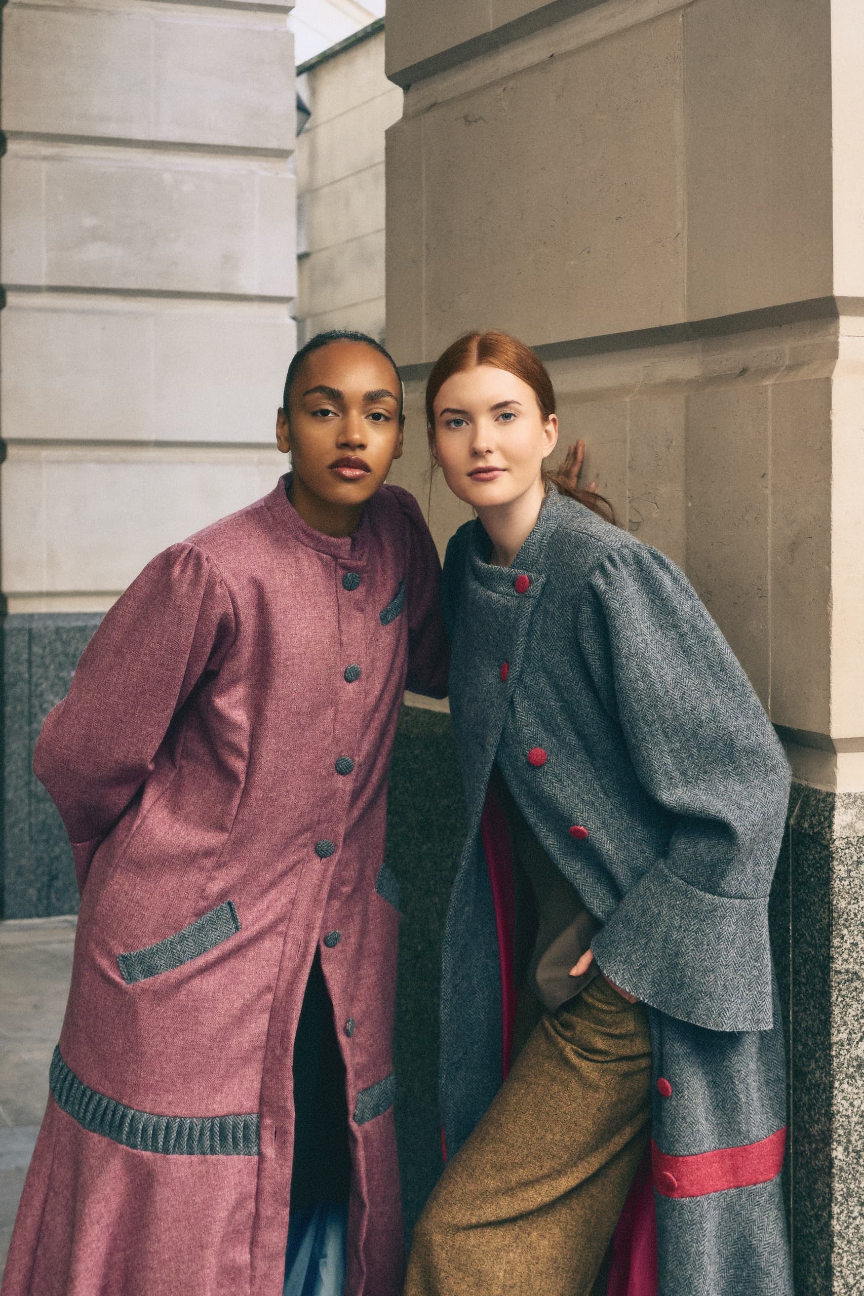 Two women in stylish overcoats pose outside an urban building.