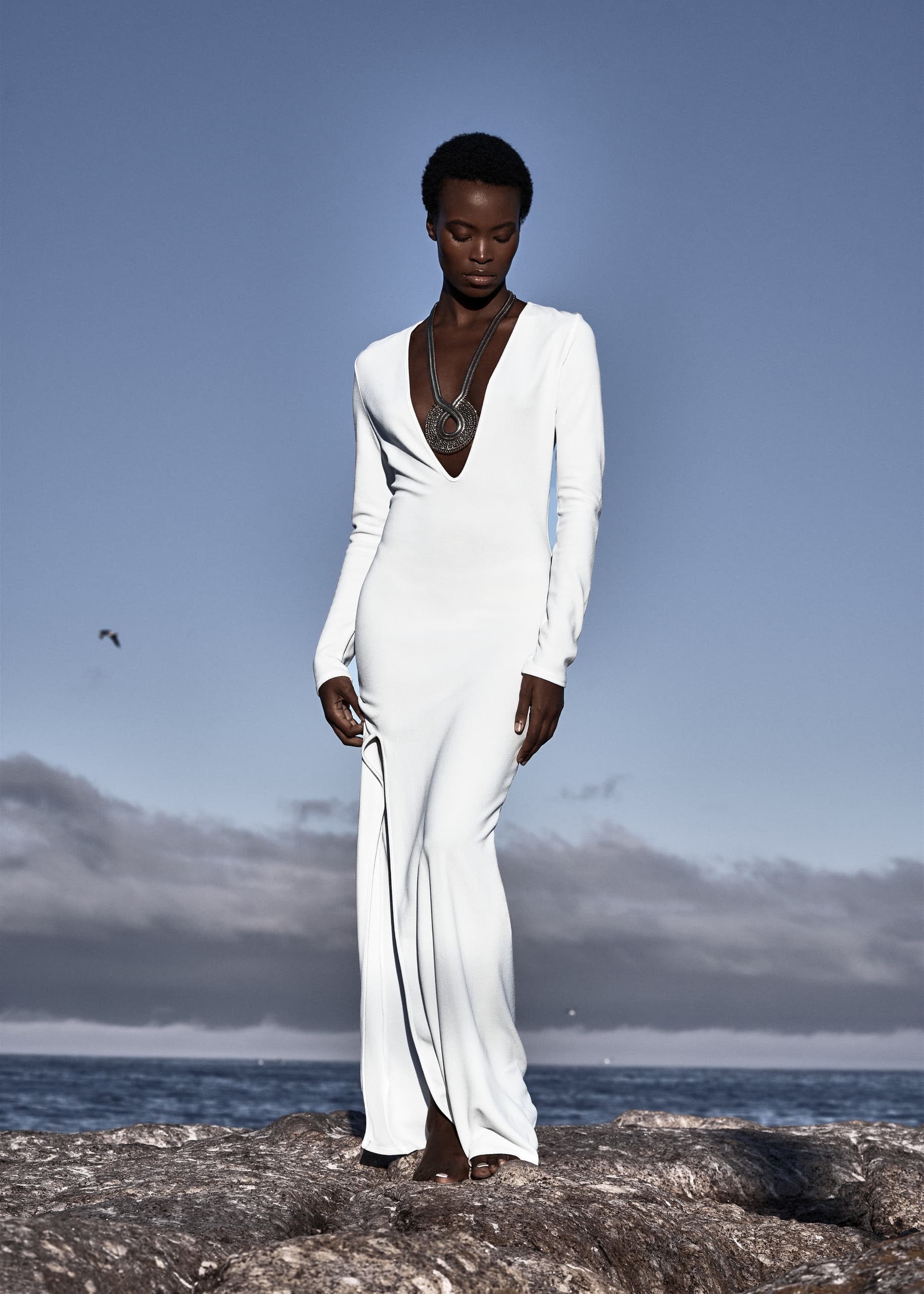 Elegant woman in white dress stands on rocky shore under clear sky.