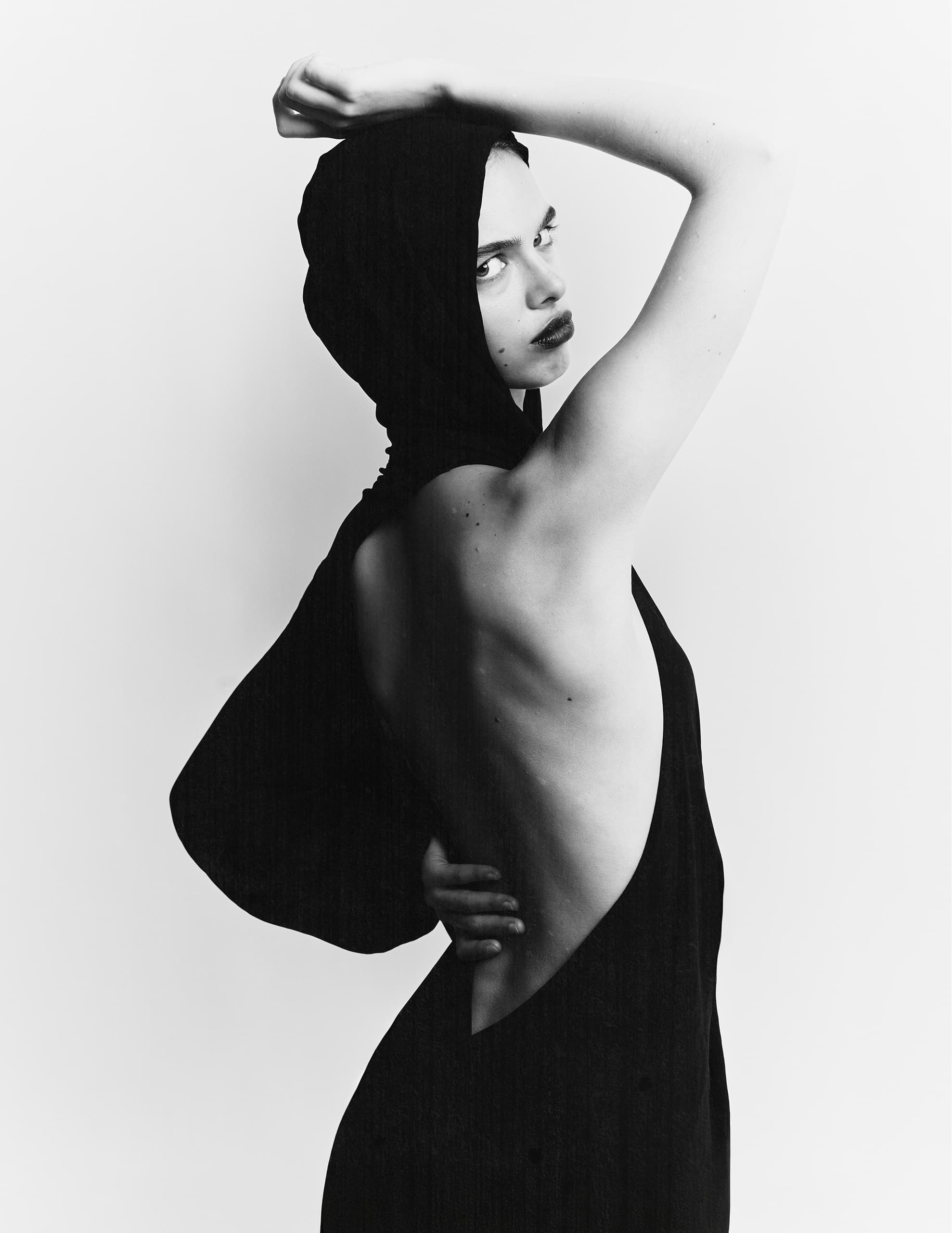 Fashion portrait of person in sleek black dress with hood, looking over their shoulder.