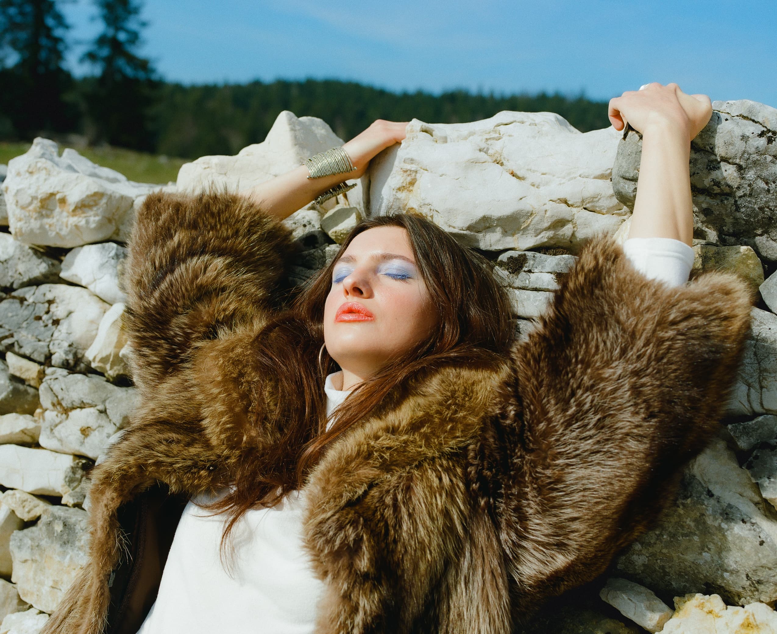 Woman in fur coat relaxing against stone wall with arms outstretched in sunlight.