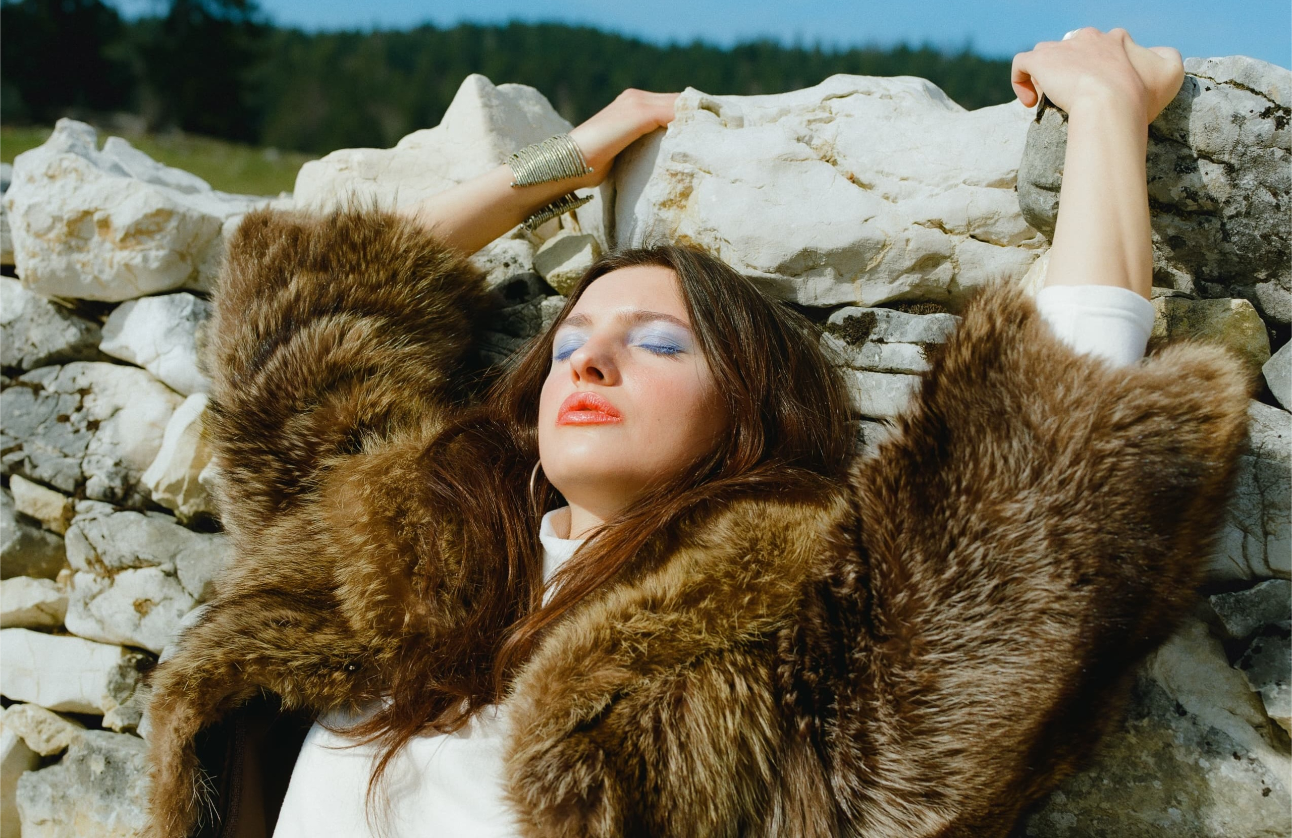 Woman in fur coat resting on a stone wall under bright sunlight.