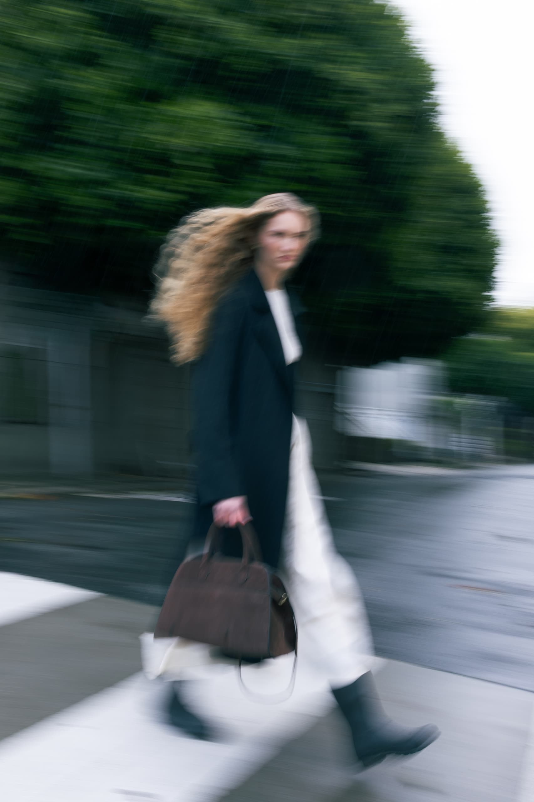 Blurry motion shot of a woman crossing a street with a handbag on a windy day.