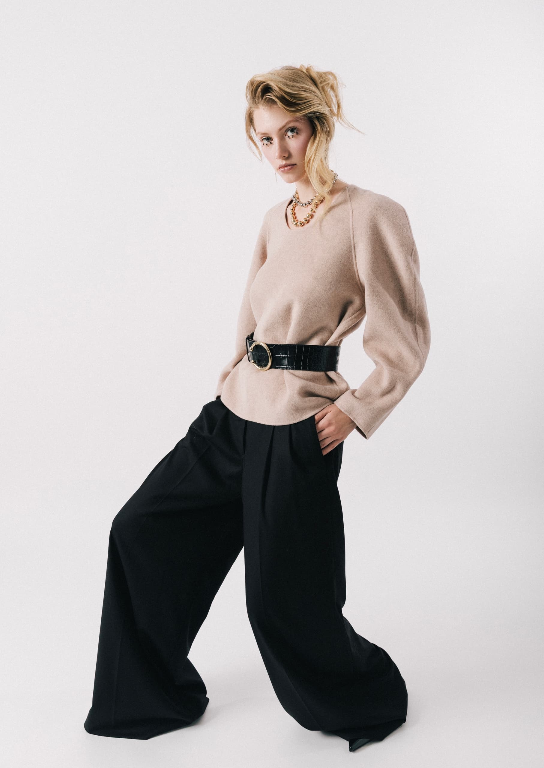 Fashionable woman in beige top, black pants, stylish pose on minimalist white background.