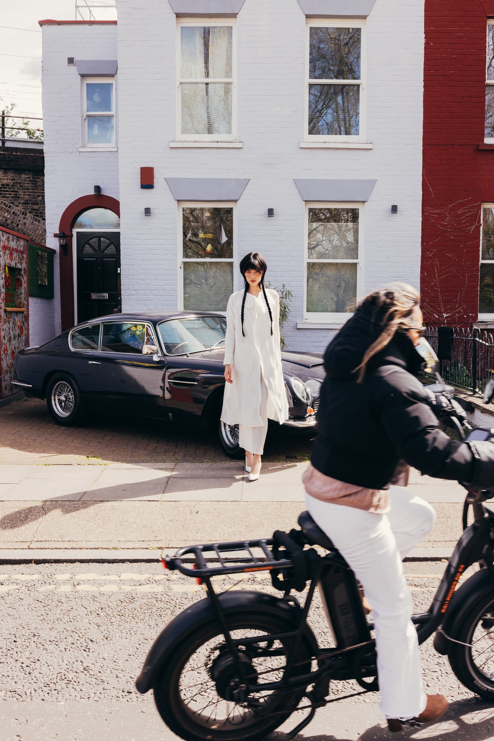 Woman in white stands near vintage car; cyclist passes by in urban street scene.