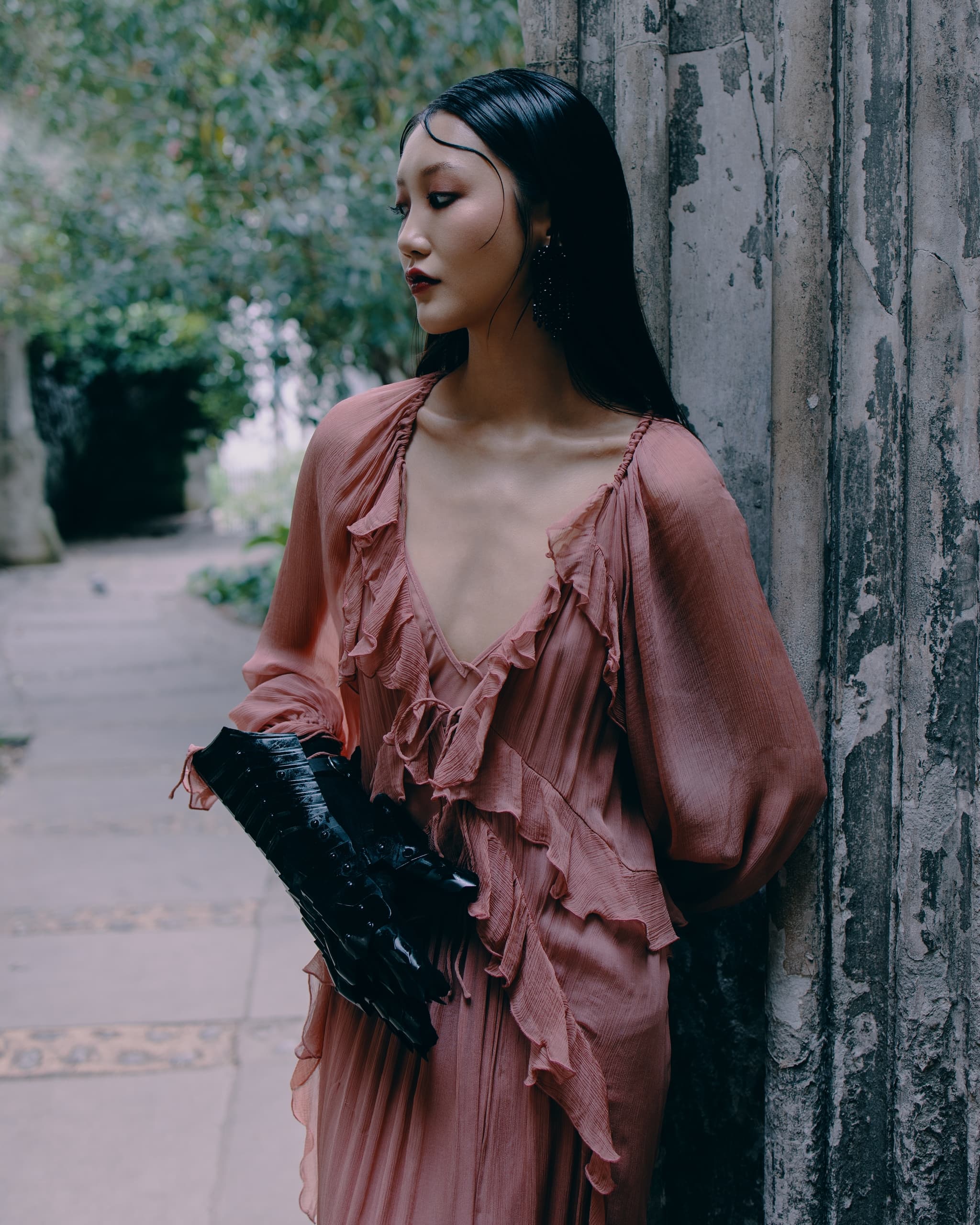 Woman in elegant pink dress and gloves, leaning against textured wall in garden.