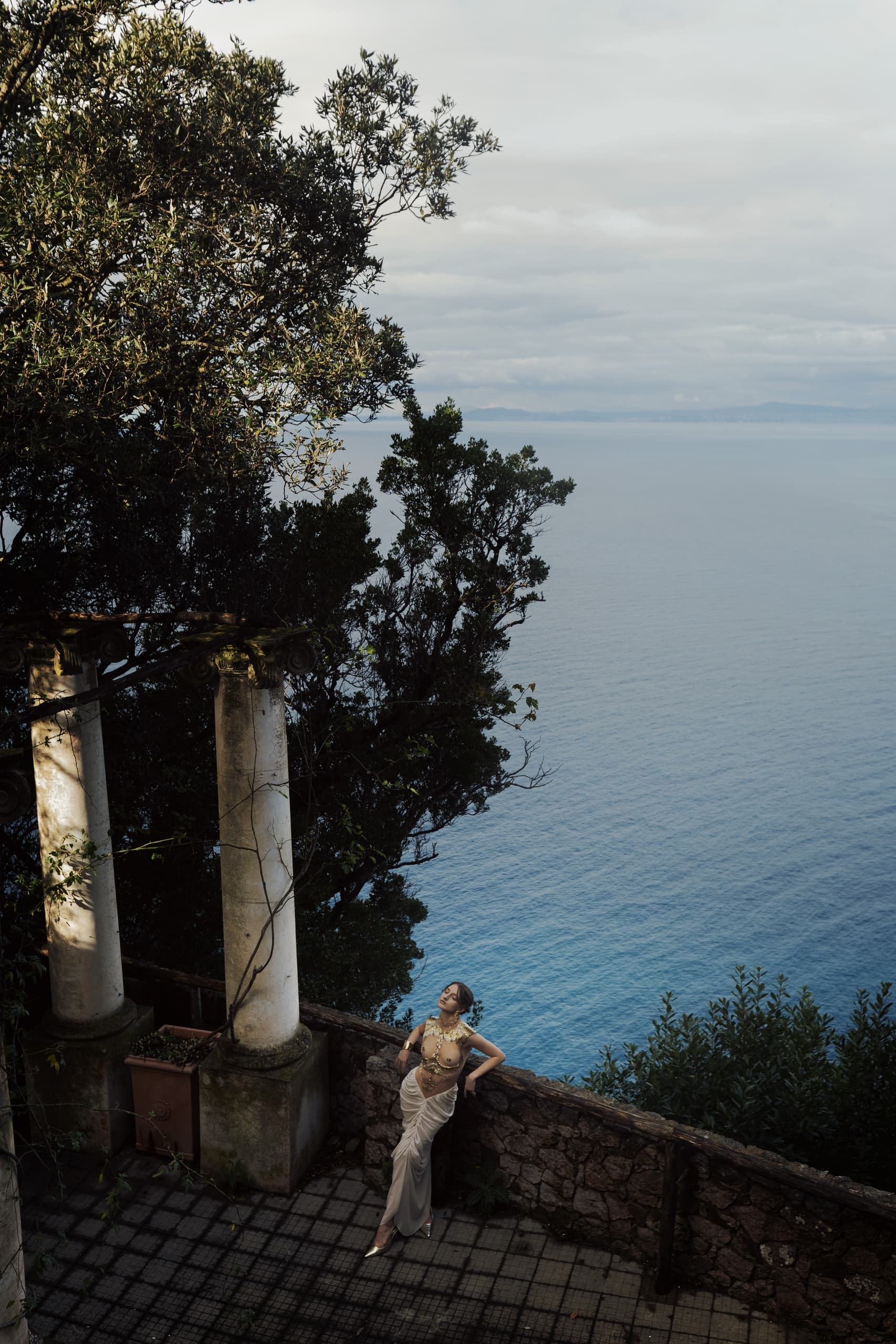 Elegant woman in gown by ocean vista with ancient columns.