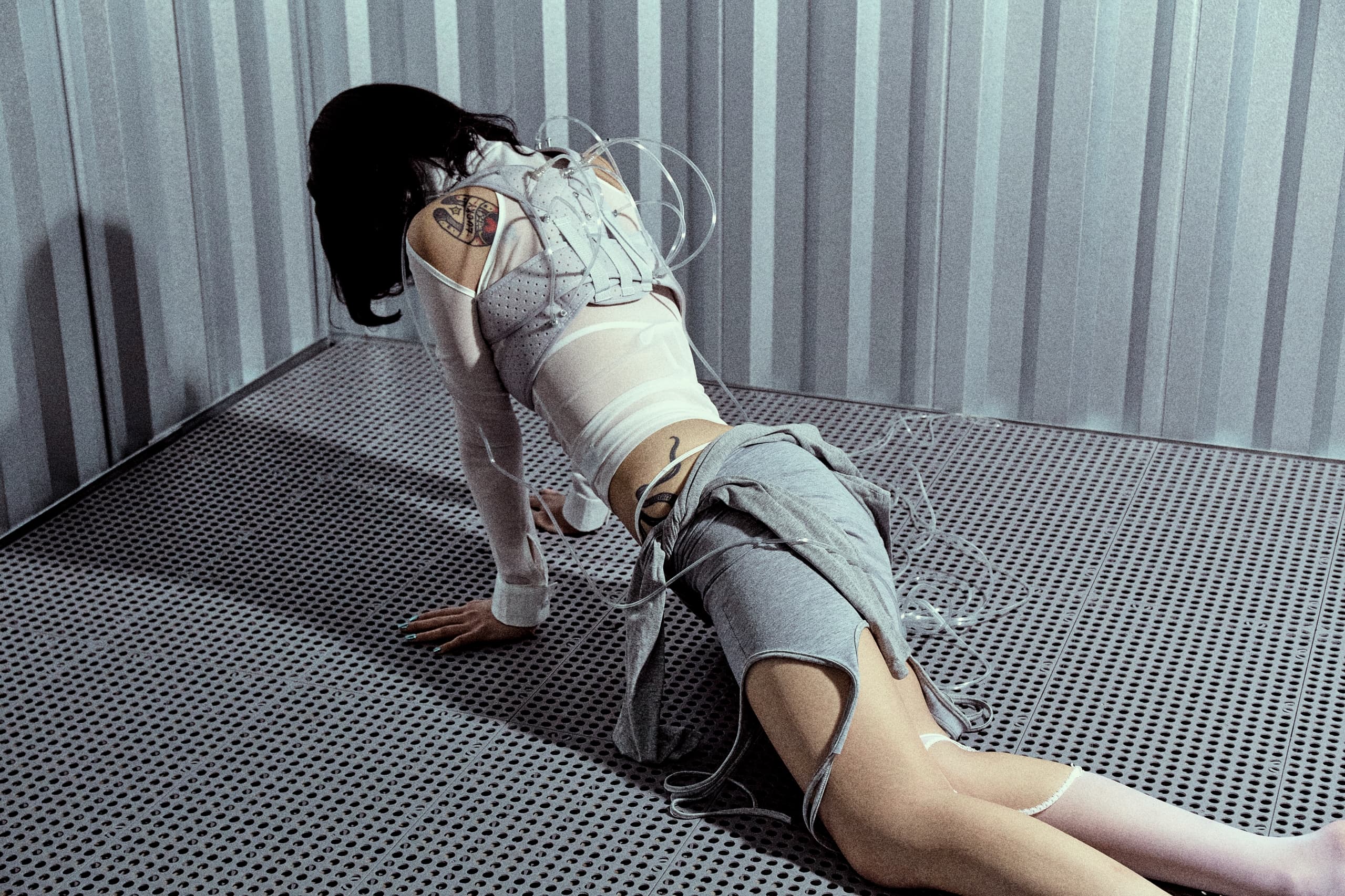 Person in futuristic attire kneels on metallic floor in a dimly lit industrial setting.