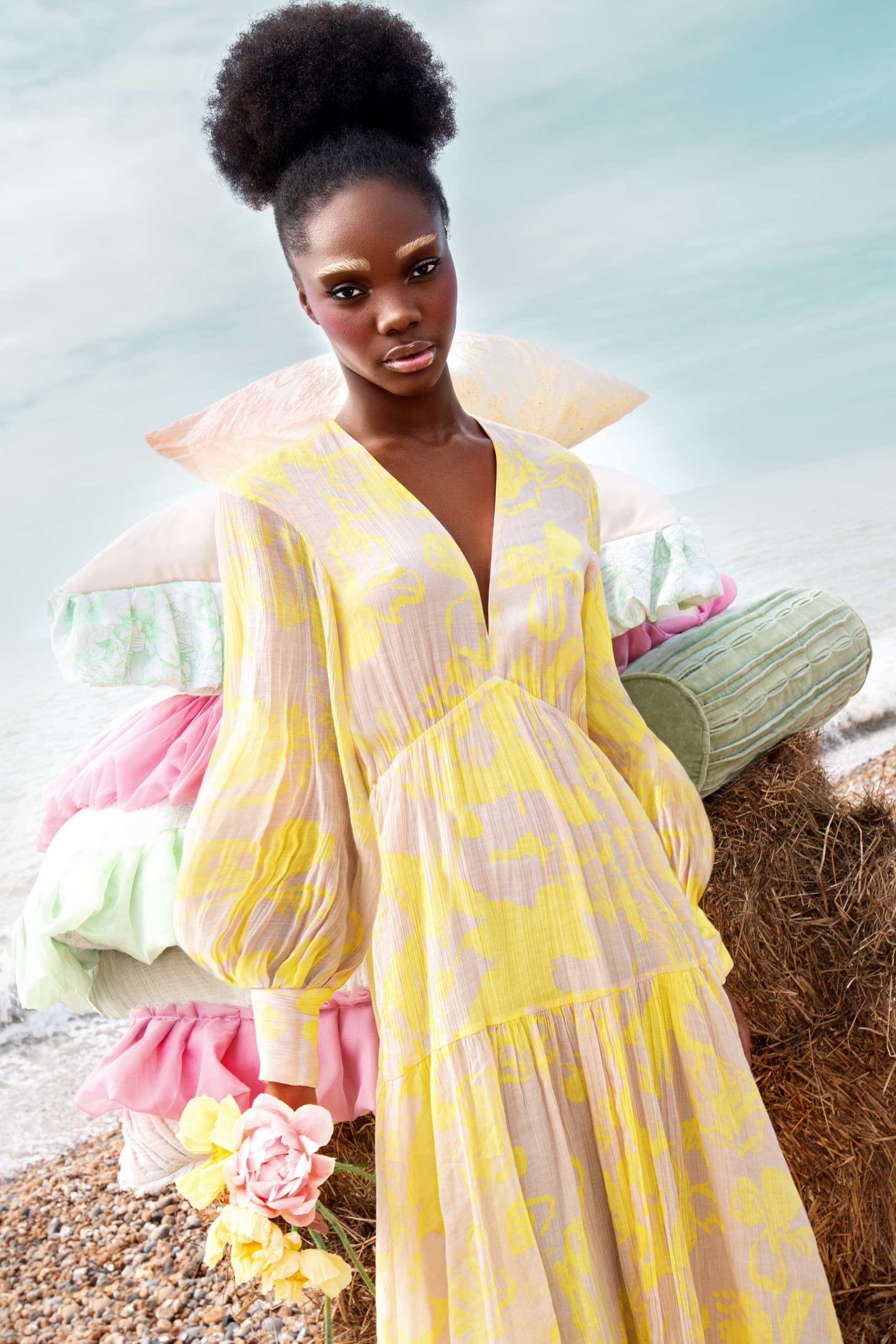 Fashion model in a yellow floral dress holding flowers on a beach with pillows.