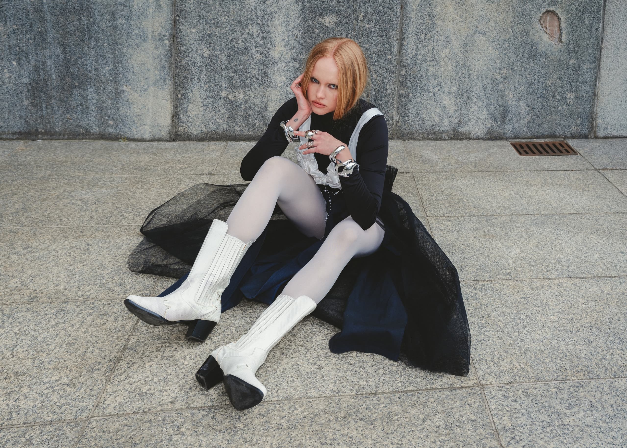 Fashionable woman in white boots sits on pavement, posing stylishly with attitude.