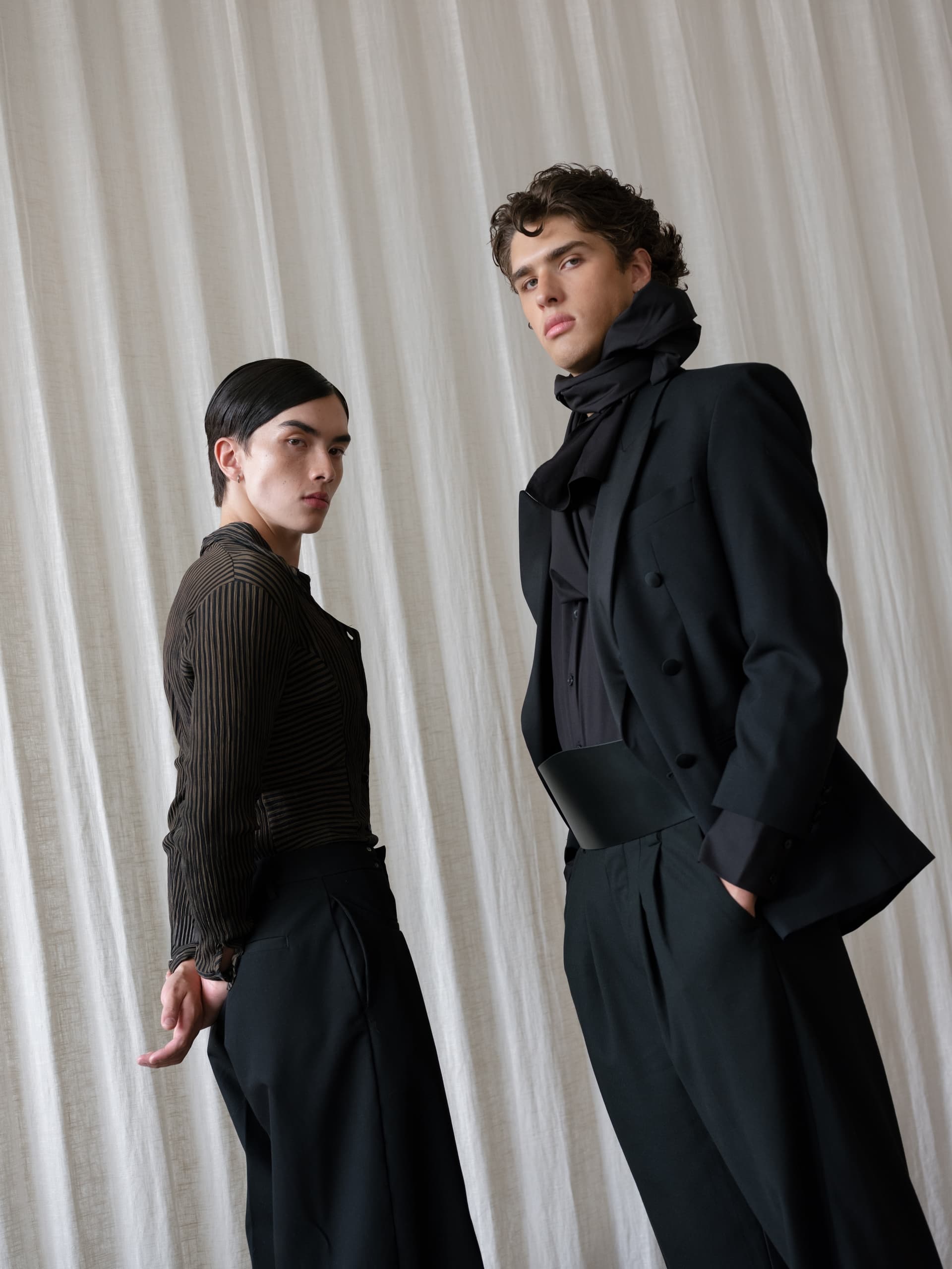 Two models in stylish outfits pose against a neutral draped curtain background.