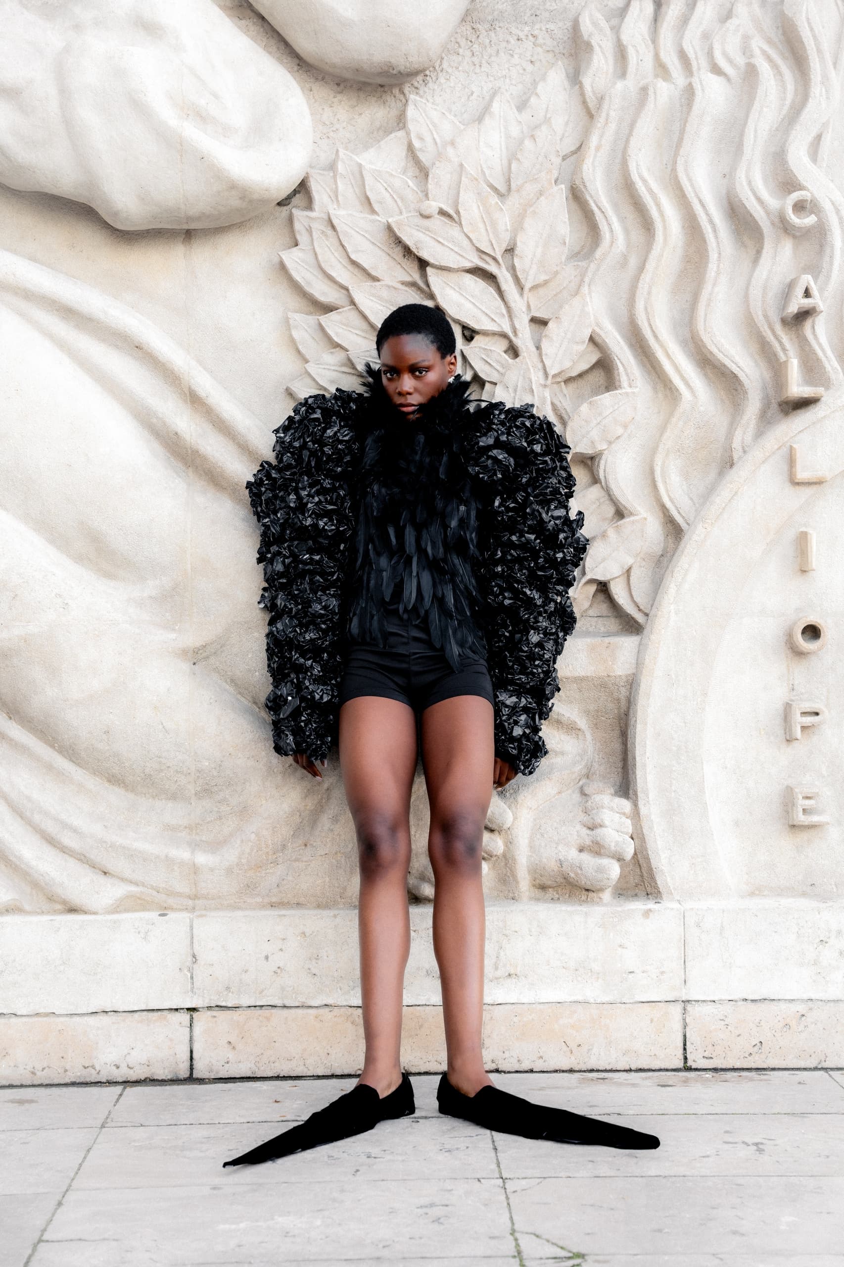 Fashion model in black textured outfit against ornate white stone background.