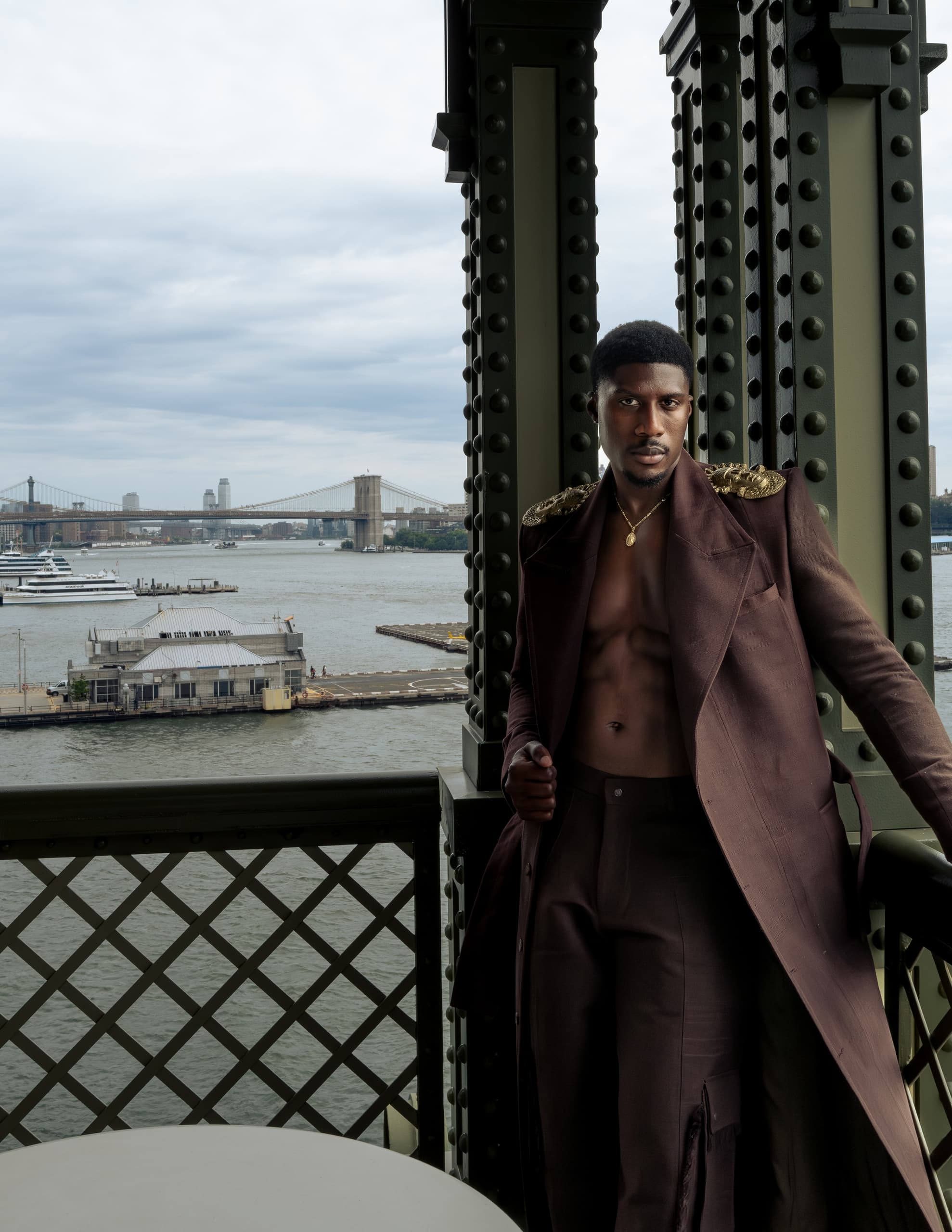 Man in stylish coat poses by riverside with cityscape and bridge in the background.