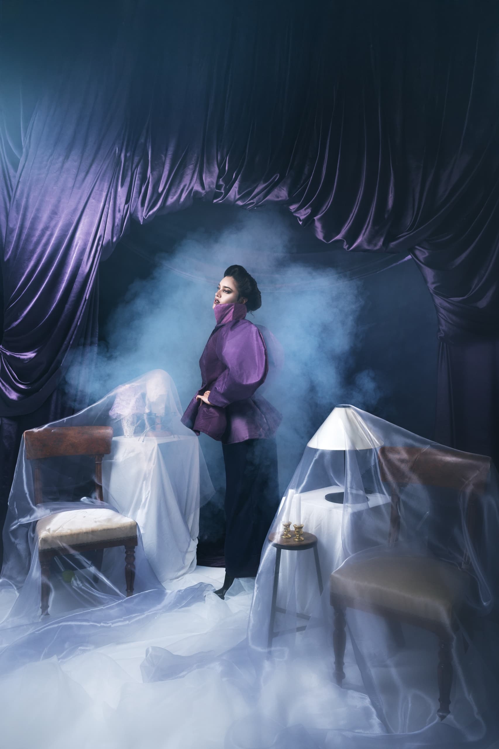 Elegant woman in purple amidst fog, draped velvet setting, moody atmosphere.