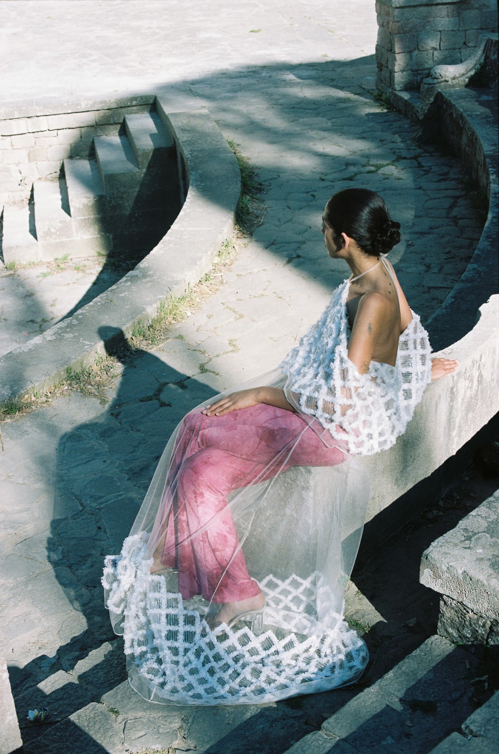 Woman in artistic, sheer white attire, sitting in a sunlit, stone stair setting.