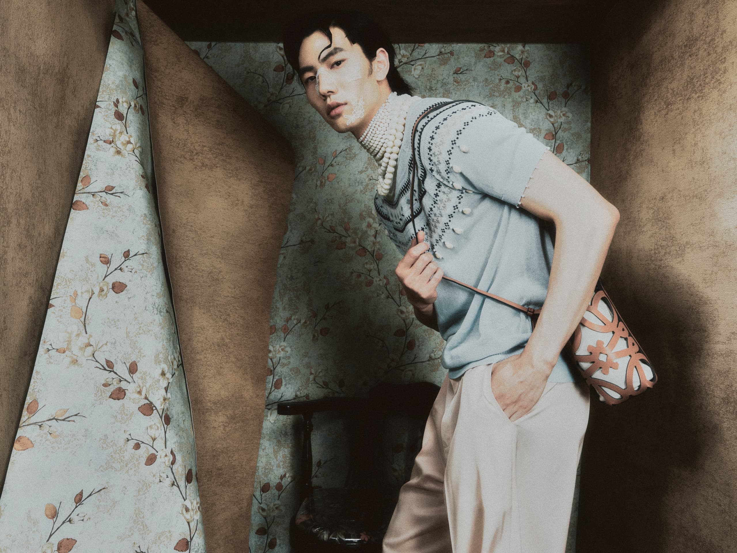 Fashionable man posing with floral wallpaper backdrop and stylish crossbody bag.