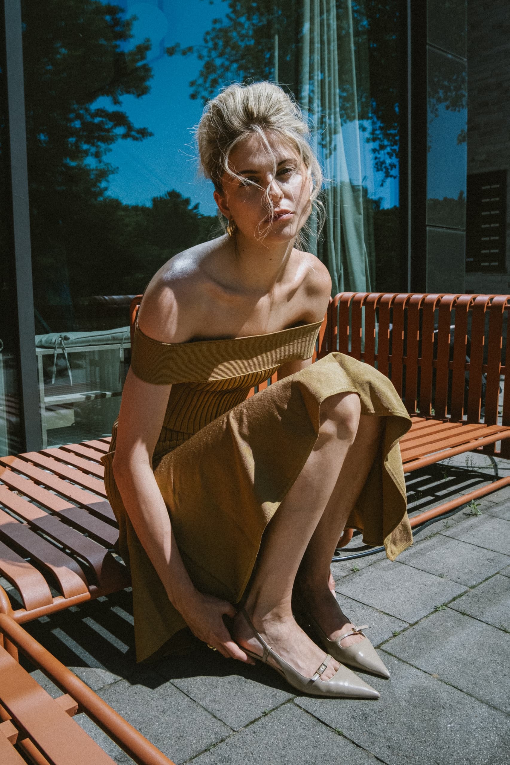 Woman in stylish brown dress squats on orange bench, outdoor sunny setting.