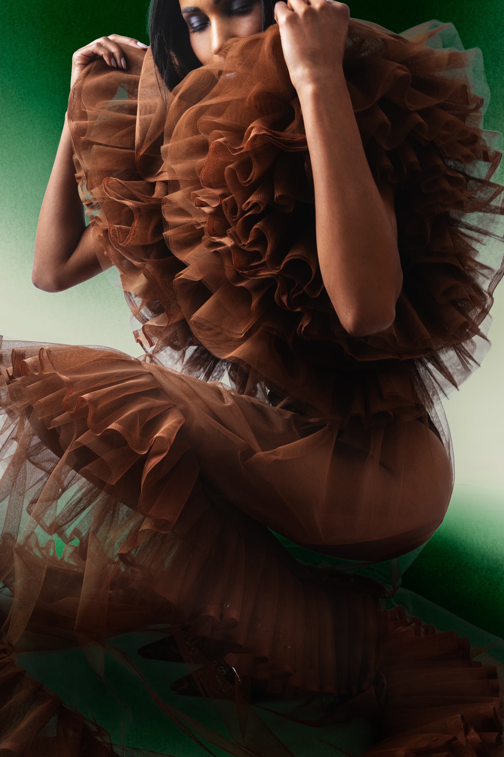 Model in brown tulle dress with dramatic ruffles, set against a green gradient background.