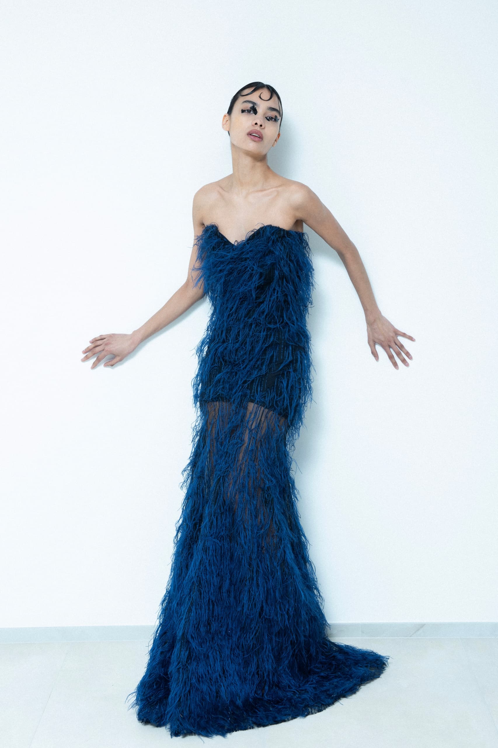 Woman in blue feathered evening gown posing elegantly against a white wall.
