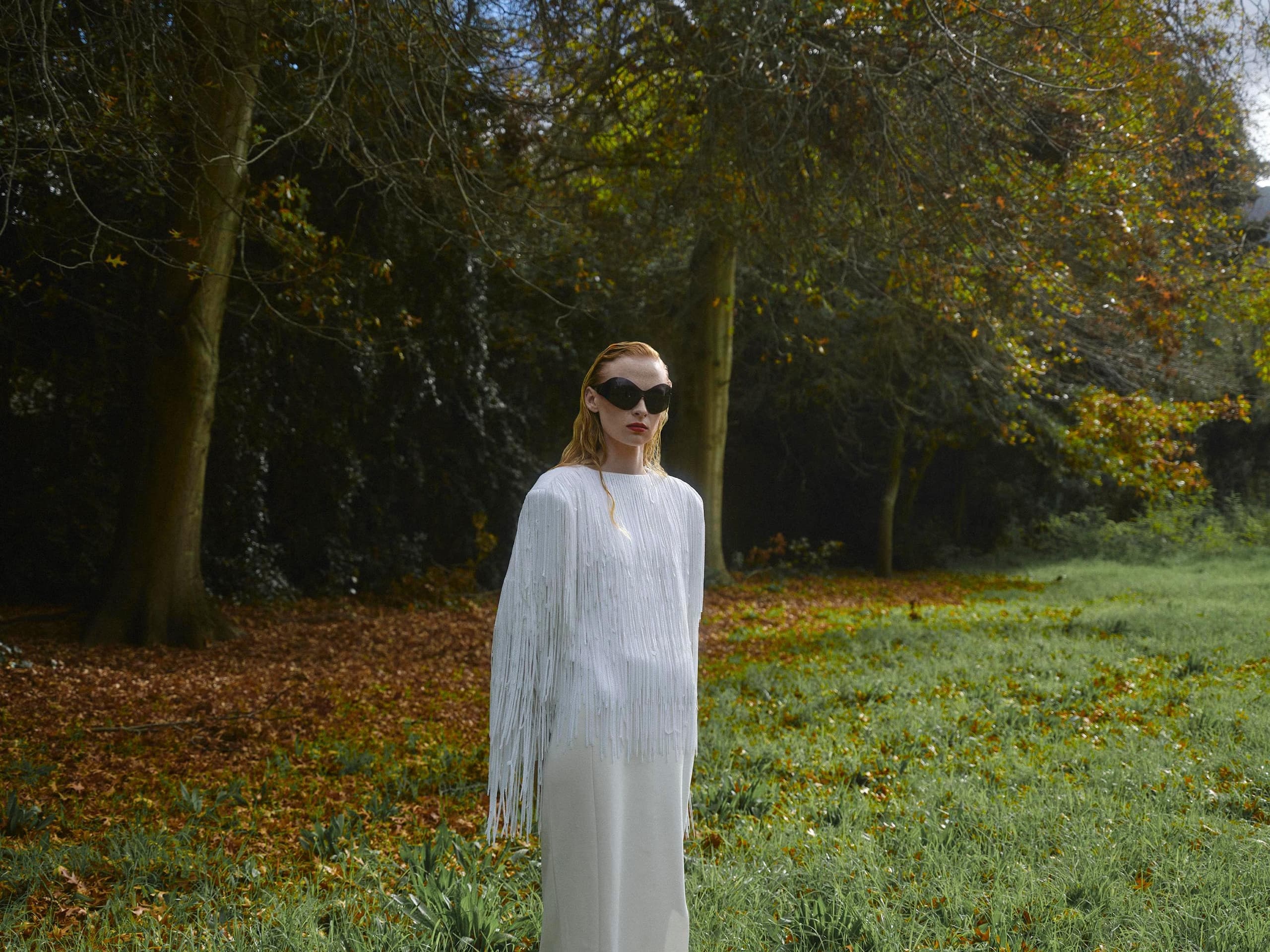 Woman in a white fringe dress with sunglasses standing in an autumnal park.